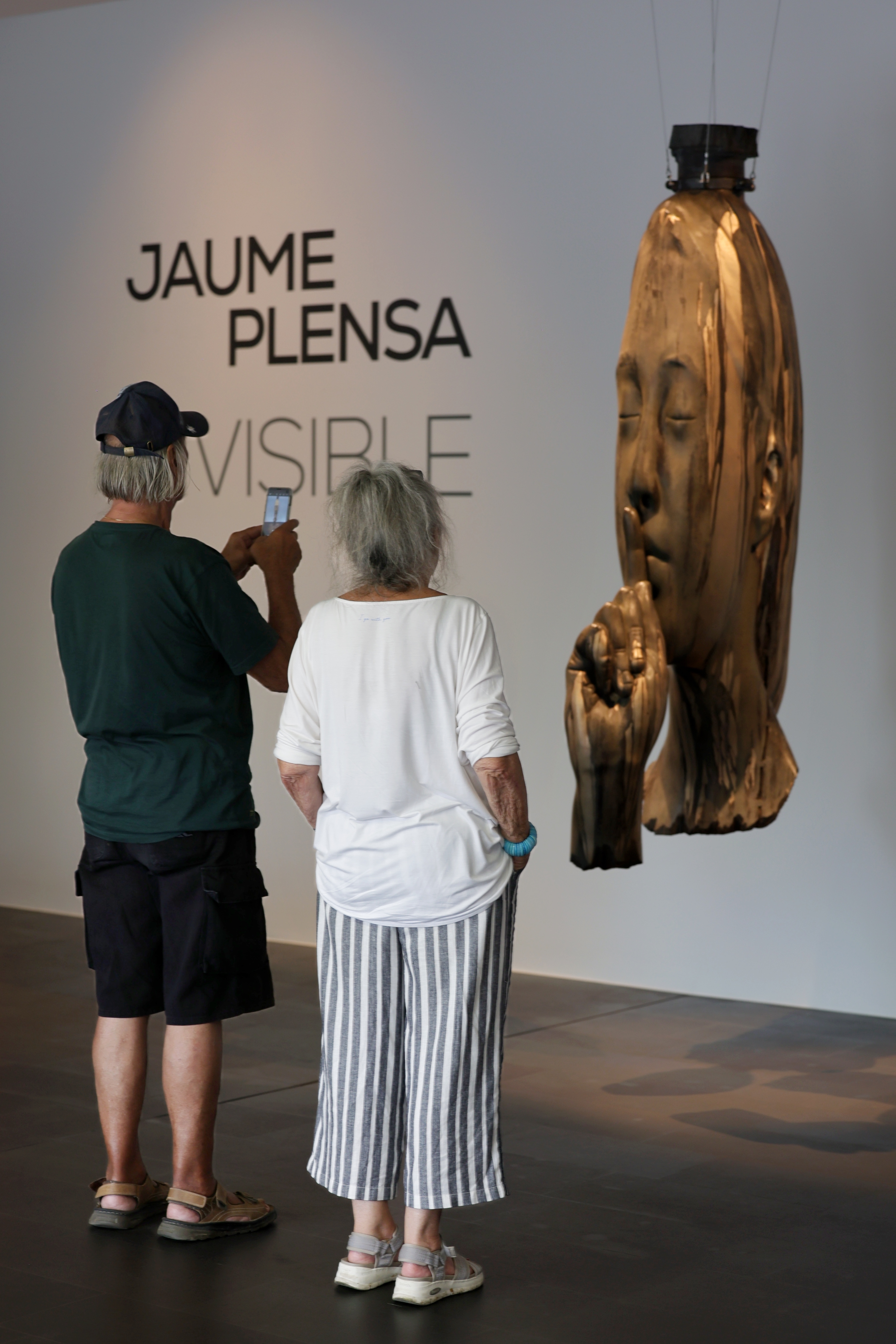 Zwei Besucher betrachten eine hängende Kopfskulptur vor dem Schriftzug Jaume Plensa Invisible im Museum Küppersmühle