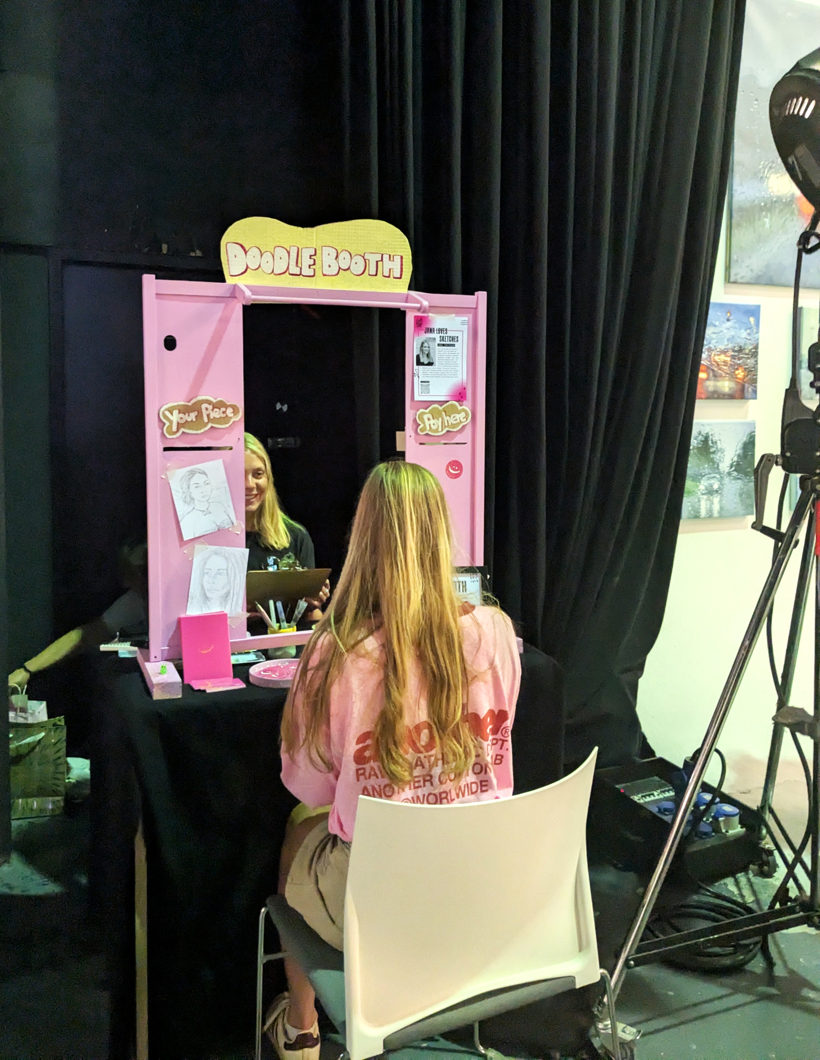 Eine Person sitzt vor einem rosa Doodle Booth, in dem eine Zeichnerin ein Porträt anfertigt