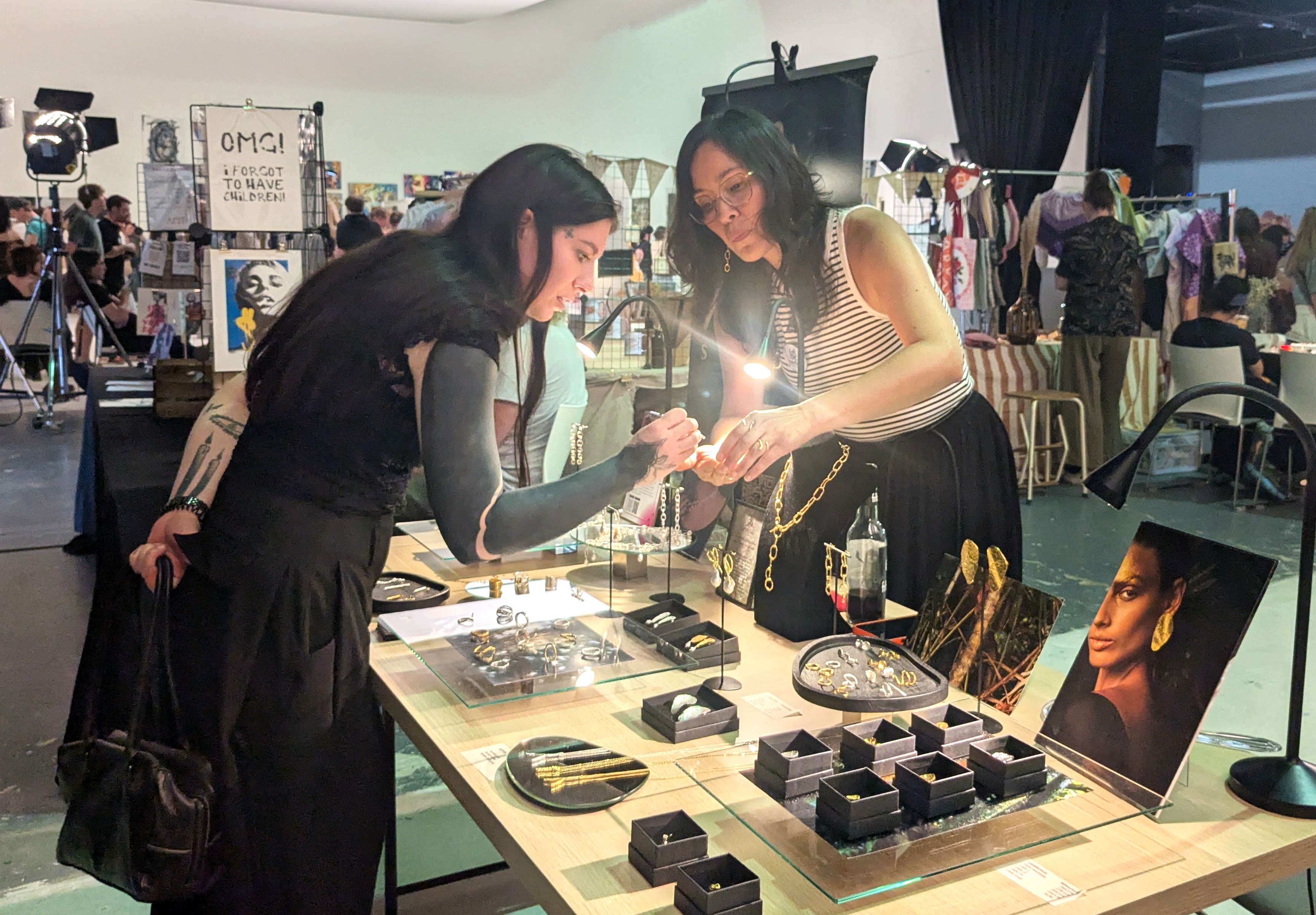 Zwei Frauen betrachten unter einer Lampe Schmuck an einem Stand, rechts ein Porträtfoto mit goldenem Blattschmuck