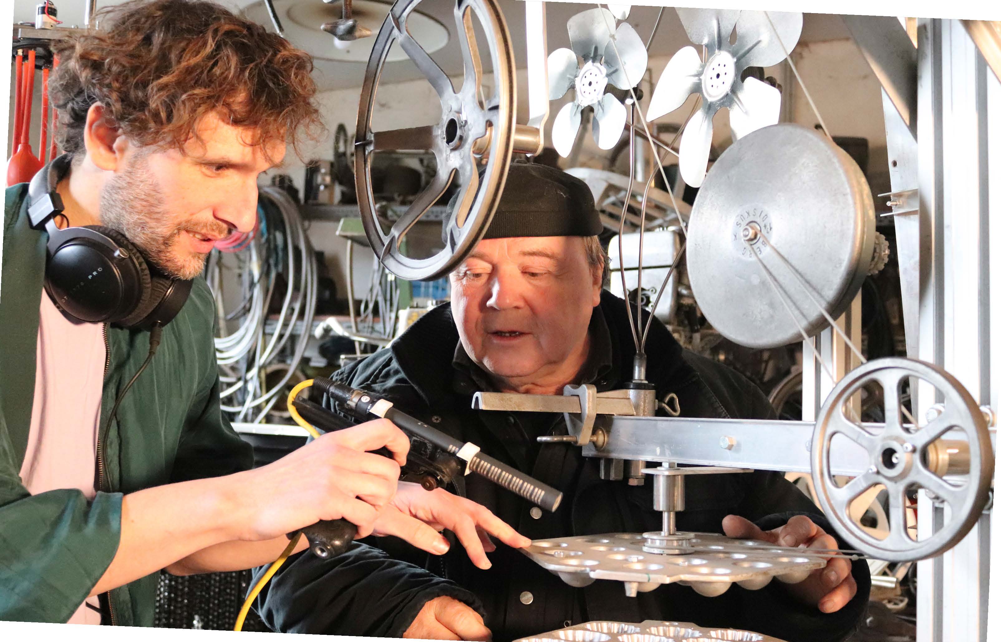 Nils Mosh und Willi Reiche arbeiten gemeinsam an einer Kunstmaschine. Mosh hält ein Mikrofon an die mechanischen Elemente, Reiche steht daneben.