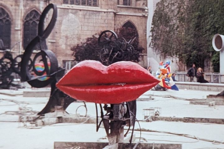 Rotes Lippenpaar aus Kunstharz in einem trockenen Brunnenbecken, dahinter schwarze Metallskulpturen und eine gotische Kirche.