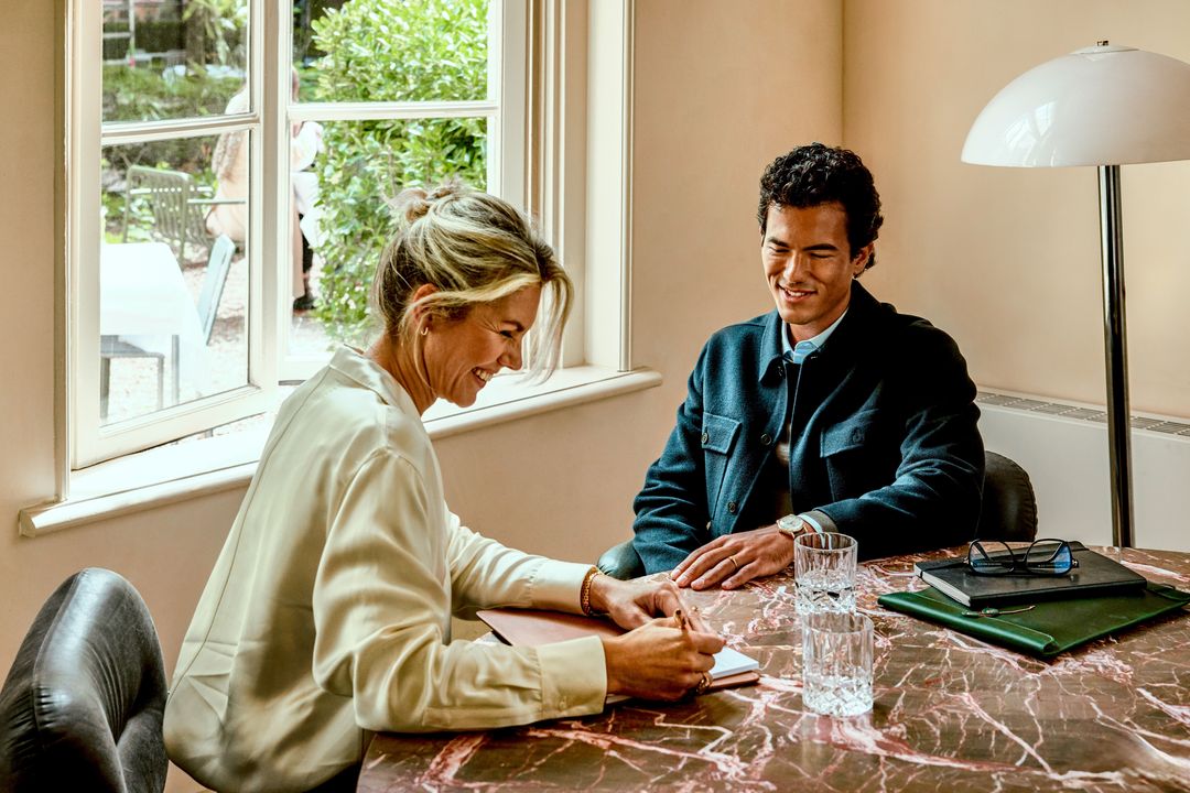 Zwei Personen im Gespräch am Tisch, eine Person macht Notizen.