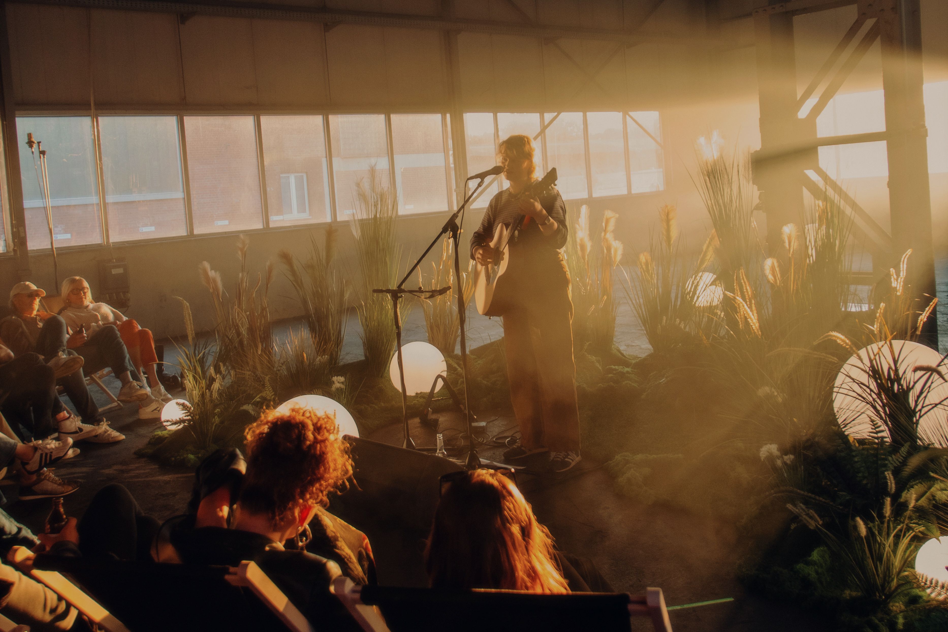 Eine Musikerin mit Gitarre singt beim Musikgarten vor Publikum in einer begrünten Industriehalle auf dem Areal Böhler. Warmes Abendlicht fällt durch die Fenster auf die Bühne.