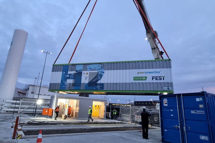 Kran hebt einen Container-Elektrolyseur („greenpowerbox“, Stadtwerke Düsseldorf/FEST) am Höherweg in Düsseldorf-Flingern über eine Baustelle; Mitarbeitende in Warnwesten stehen darunter.
