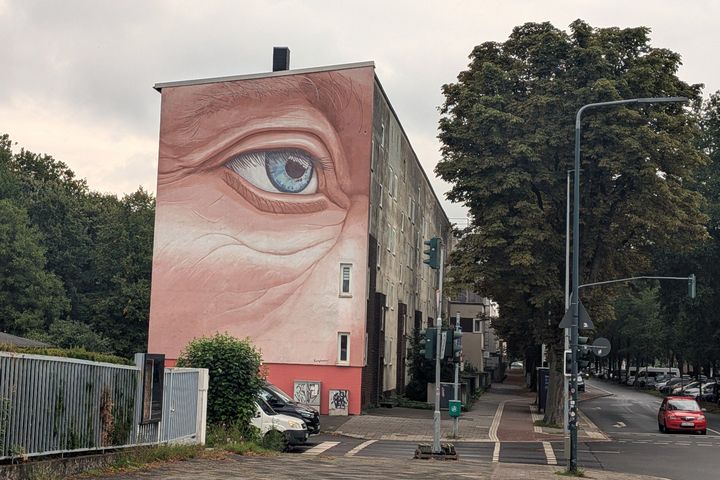 Wandbild „Auge“ am Hellweg in Düsseldorf-Flingern Das Wandbild „Auge“ an der rosafarbenen Giebelwand eines Wohnhauses am Hellweg in Düsseldorf-Flingern, davor eine Straßenkreuzung mit Ampeln und Bäumen