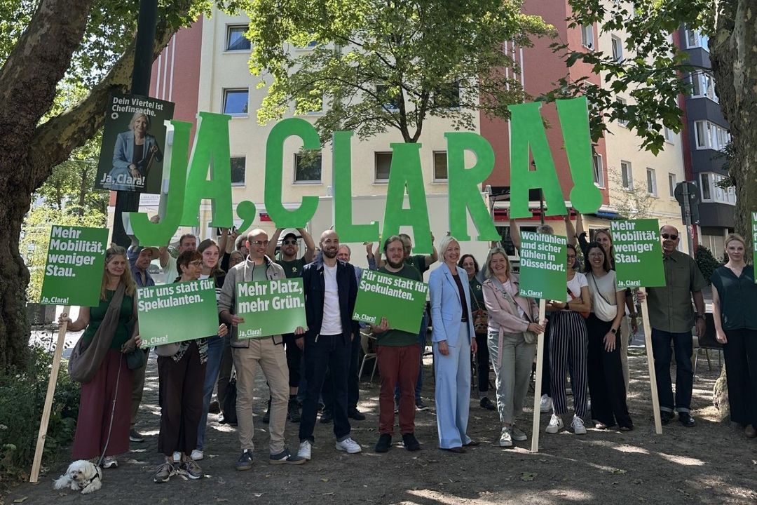Wahlkampfauftakt mit Botschaften aus den Stadtteilen: Clara Gerlach (Grüne, vorne rechts im hellblauen Anzug) präsentierte am 7. August 2025 auf dem Maria-und-Josef-Otten-Platz ihr Sofortprogramm zur OB-Wahl. Unterstützer:innen hielten Schilder mit den zentralen Forderungen: mehr Grün, weniger Stau, Schutz für Mieter:innen. / Foto: Foto: BÜNDNIS 90/DIE GRÜNEN Düsseldorf
