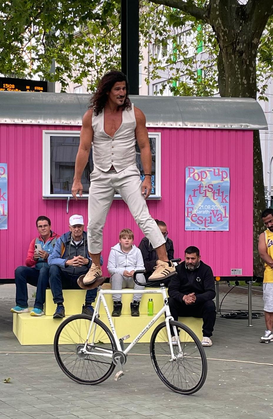 Ein Artist balanciert im öffentlichen Raum stehend auf einem Fahrrad, während Zuschauerinnen und Zuschauer die Vorstellung vor einem pinkfarbenen Festivalwagen verfolgen.