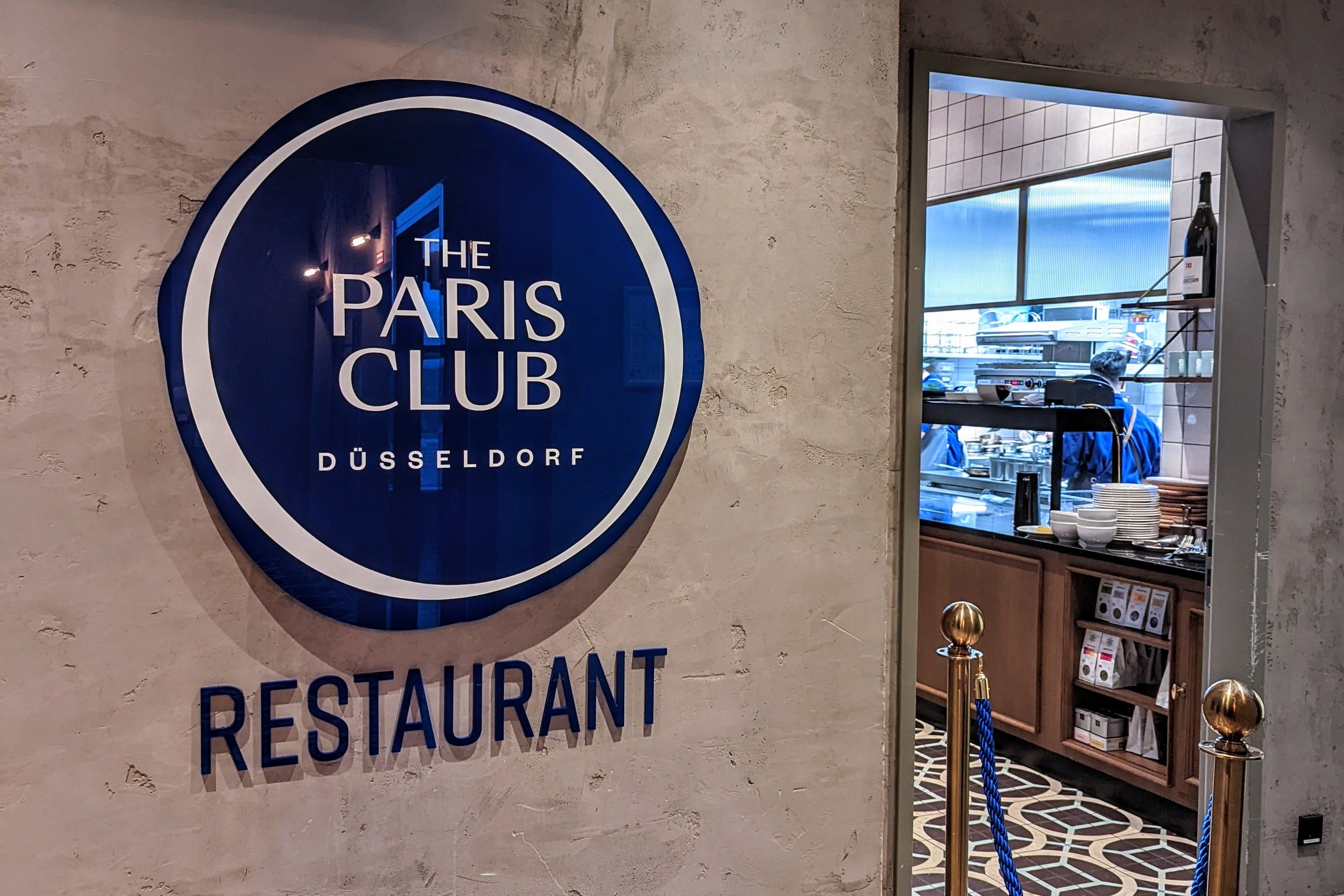 Eingang des Restaurants The Paris Club im 25hours Hotel Das Tour in Düsseldorf mit beleuchteter Logoschild und Blick in die offene Küche