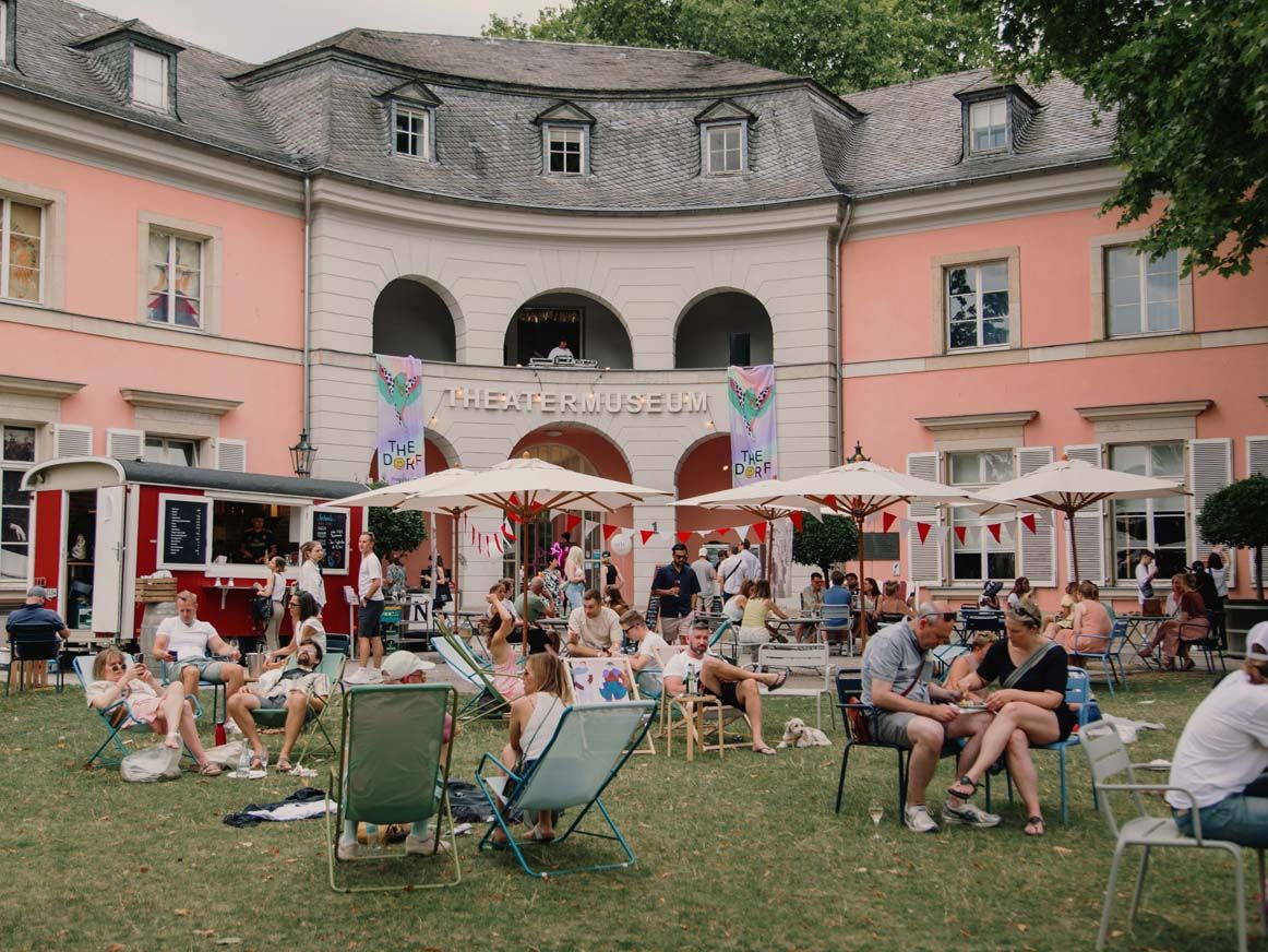 Besucherinnen und Besucher sitzen im Hofgarten vor dem Theatermuseum Düsseldorf auf Liegestühlen und Stühlen unter Sonnenschirmen.
