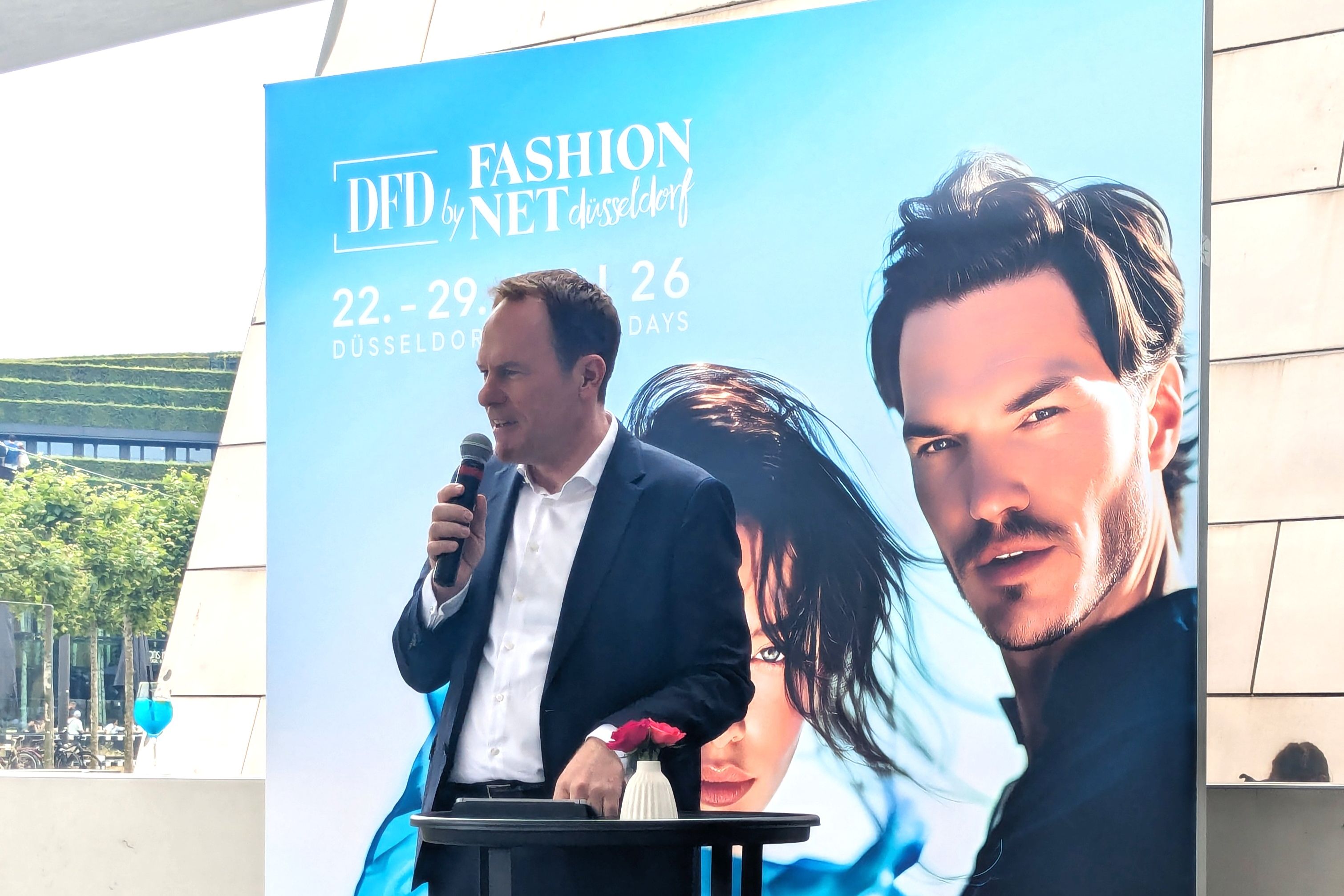 Oberbürgermeister Dr. Stephan Keller spricht in ein Mikrofon an einem schwarzen Stehpult, hinter ihm das blaue Kampagnenmotiv der DFD by Fashion Net