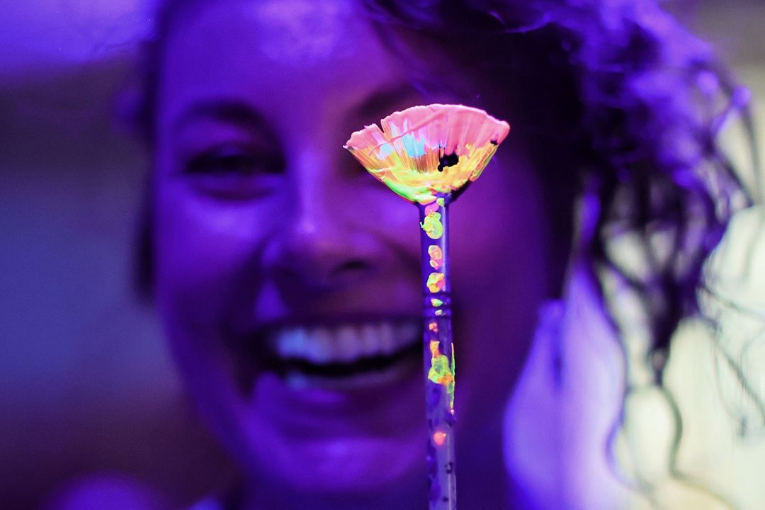 Carolin Herzberg lächelt hinter einem mit fluoreszierender Farbe bedeckten Fächerpinsel.