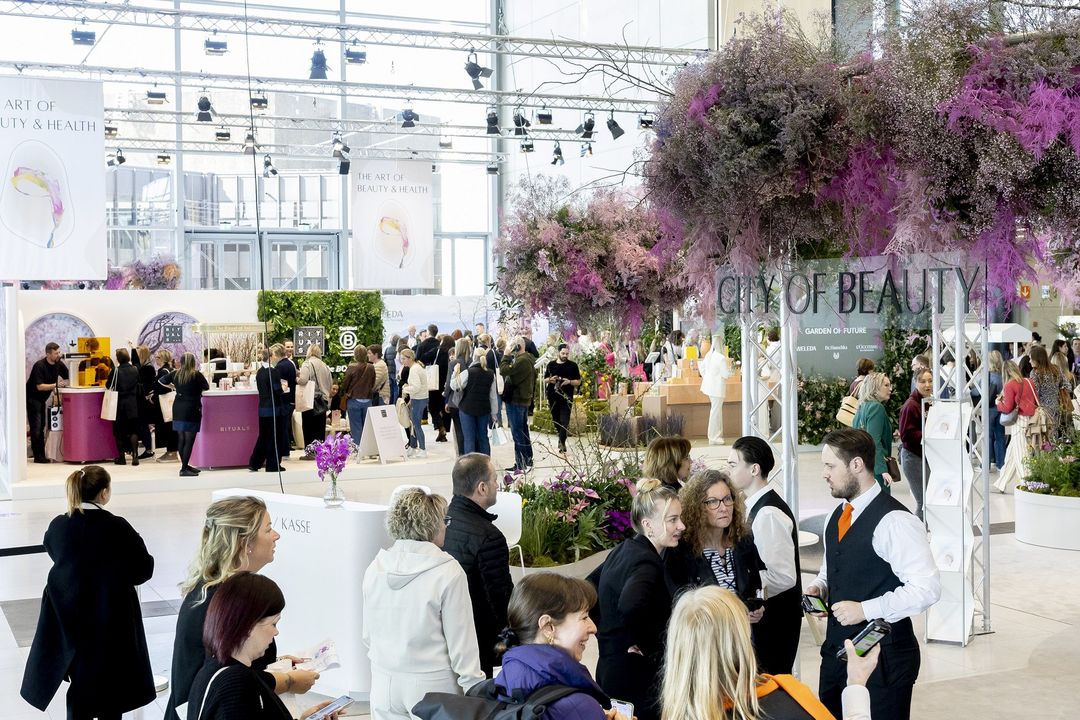 Besucherinnen und Besucher im Glasfoyer der Messe Düsseldorf bei The Art of Beauty & Health, mit floraler Dekoration und „City of Beauty“-Schriftzug.