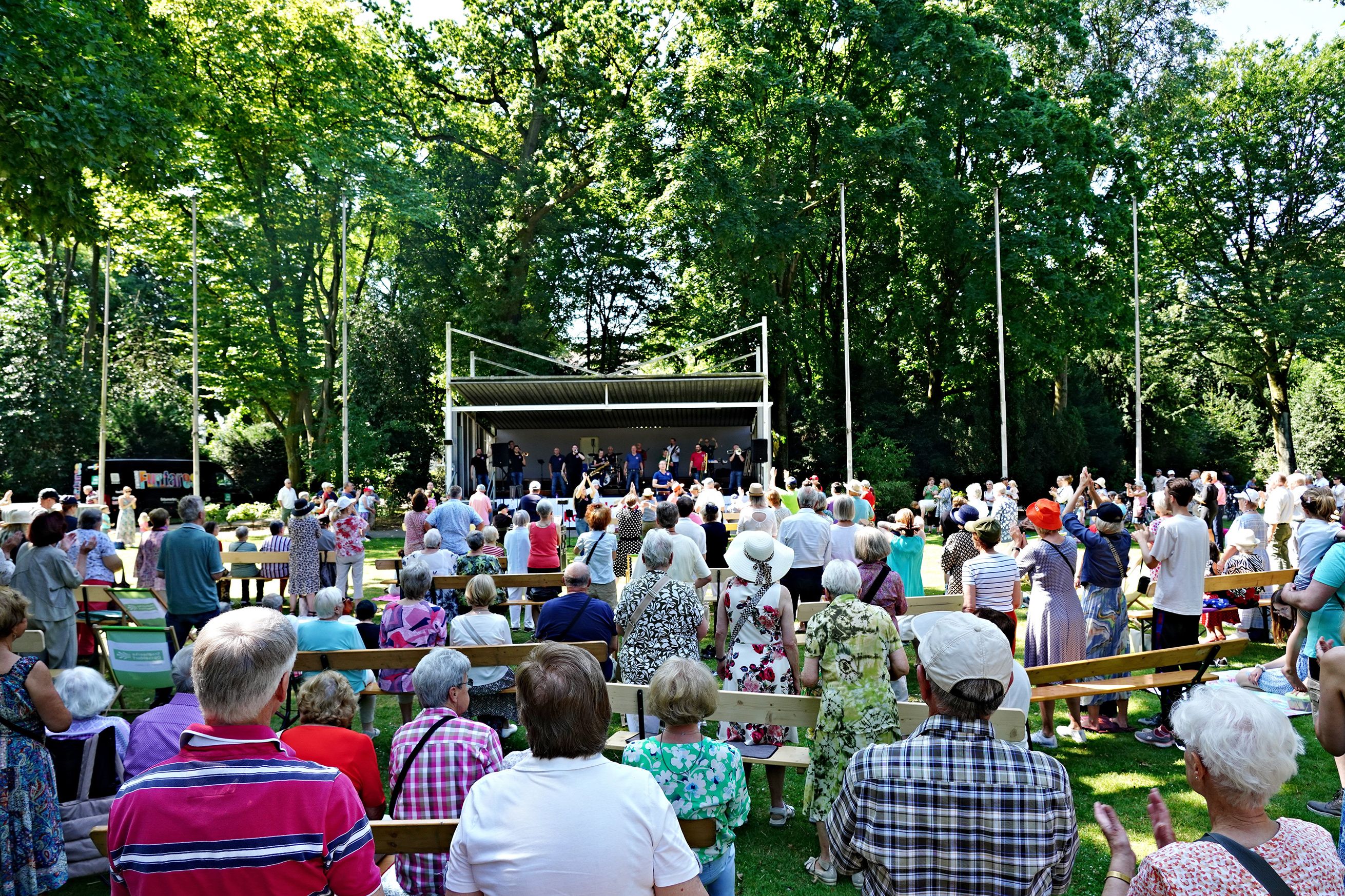 Zahlreiche Besucherinnen und Besucher sitzen und stehen vor dem Musikpavillon im Düsseldorfer Hofgarten, während eine Band auf der Bühne spielt.