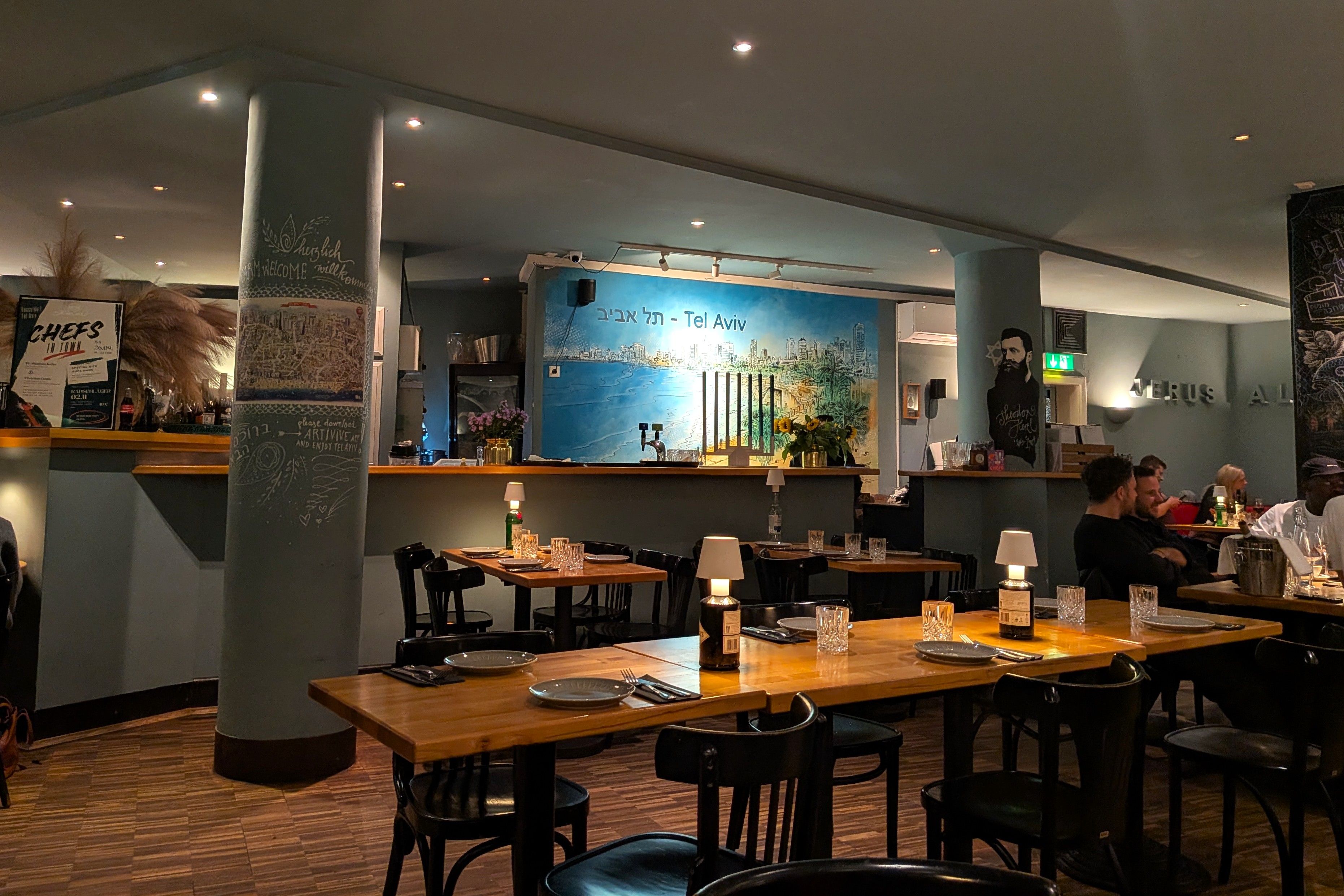 Innenraum des Restaurants Die Kurve mit Holztischen, graugrünen Wänden und großem Tel-Aviv-Wandbild.