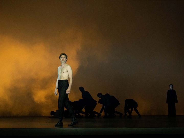 Prinz Friedrich von Homburg am Düsseldorfer Schauspielhaus André Kaczmarczyk steht mit nacktem Oberkörper vor dem gebückten Ensemble auf einer ockergelb beleuchteten, von Nebel erfüllten Bühne.