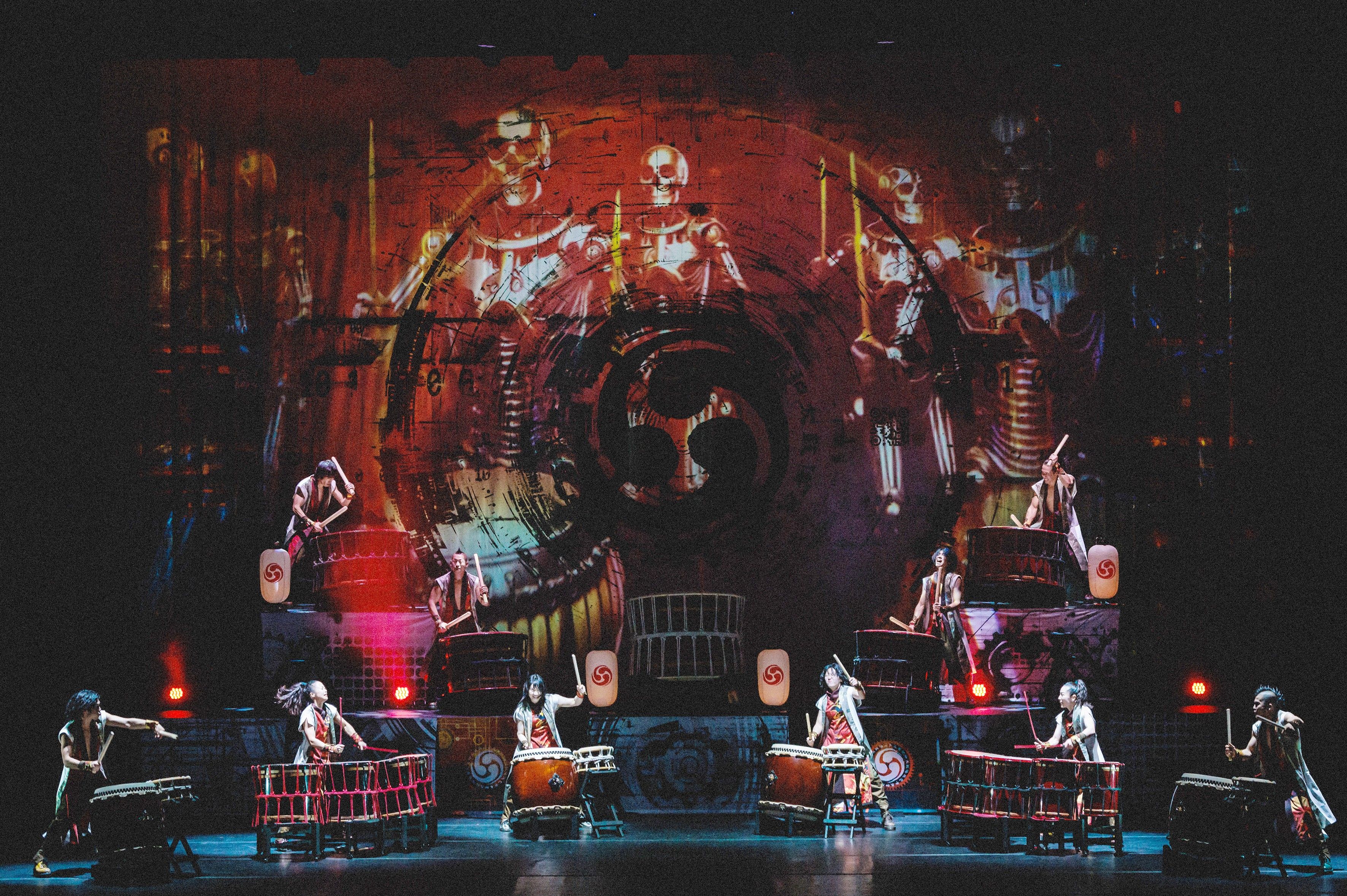 Bühnenaufnahme von YAMATO – The Drummers of Japan mit mehreren Trommlerinnen und Trommlern vor einer großflächigen Licht- und Videoprojektion.