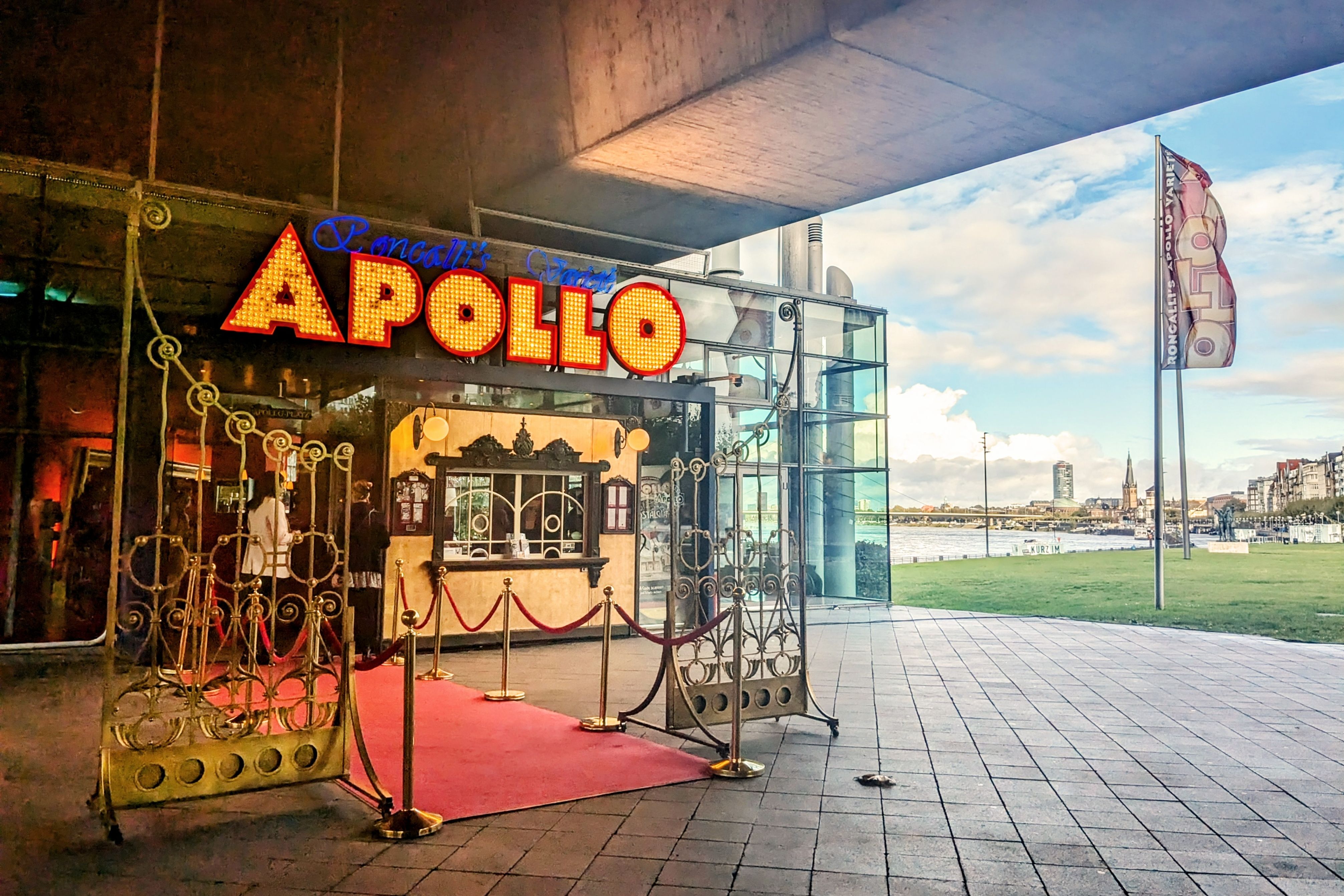 Eingang des Roncalli's Apollo Varieté in Düsseldorf mit rotem Teppich, goldenen Absperrungen und beleuchteter APOLLO-Enseigne, im Hintergrund der Rhein