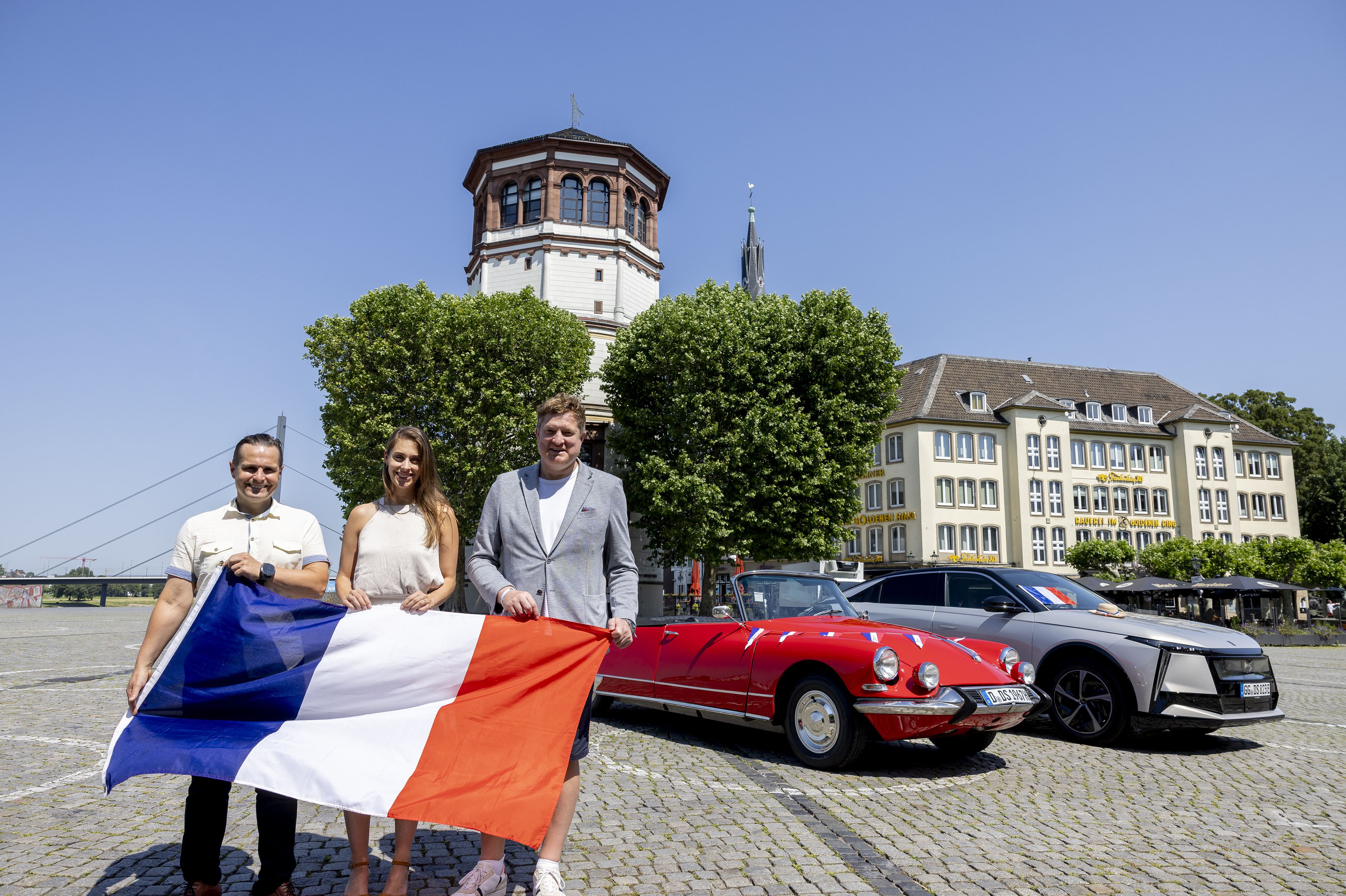 Drei Personen stehen am Düsseldorfer Rheinufer vor einem historischen roten DS-Cabrio und einem vollelektrischen DS N°8 und halten eine französische Flagge.