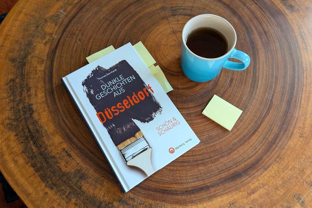 Das Buch „Dunkle Geschichten aus Düsseldorf” von Thomas Bernhardt mit gelben Haftnotizen und einer blauen Kaffeetasse auf einem Holztisch.