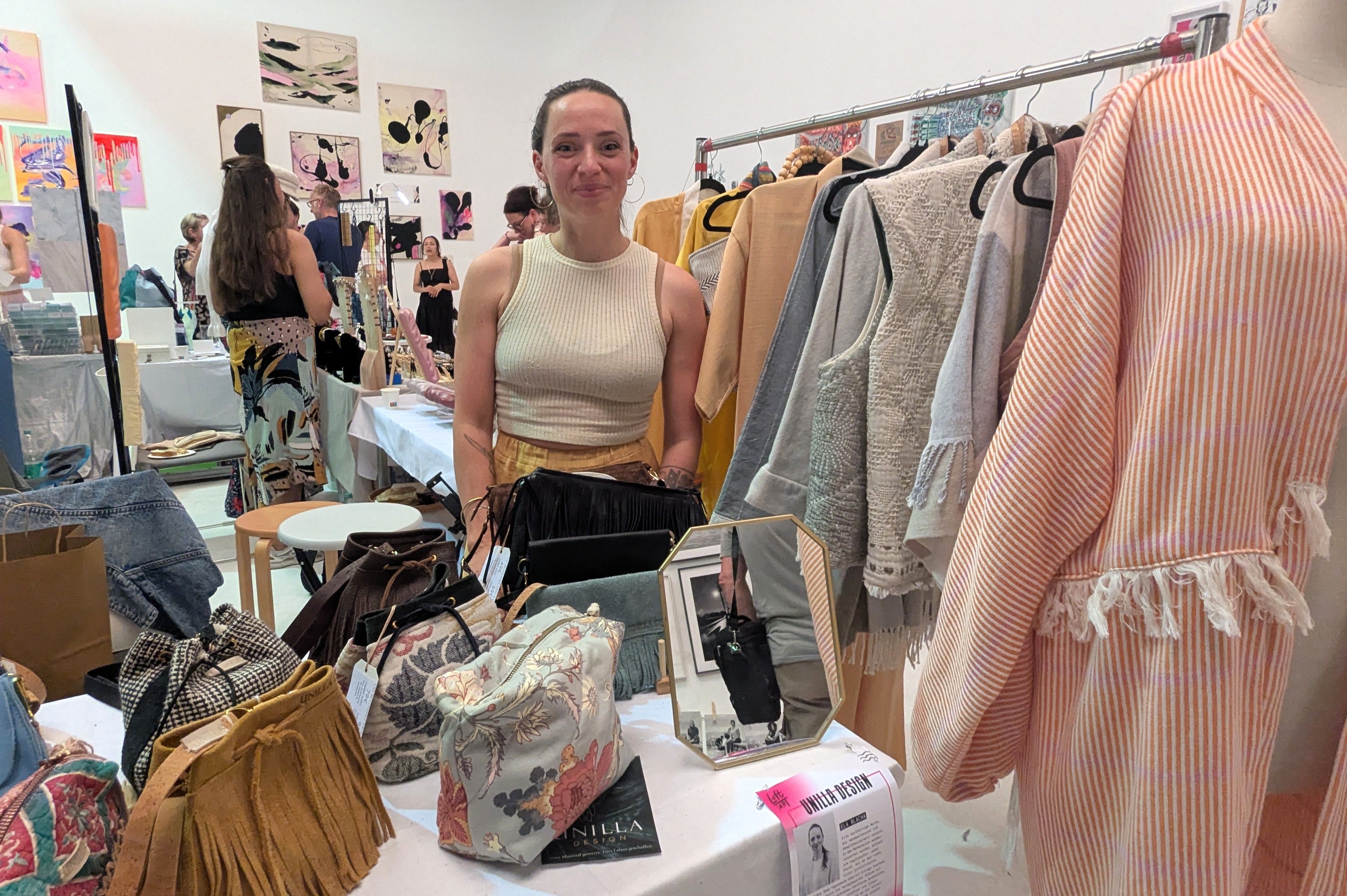 Eine Frau steht an einem Stand mit handgemachten Taschen, dahinter eine Kleiderstange mit hellen Kimonos und ärmellosen Westen, vorne ein Schild Unilla Design.
