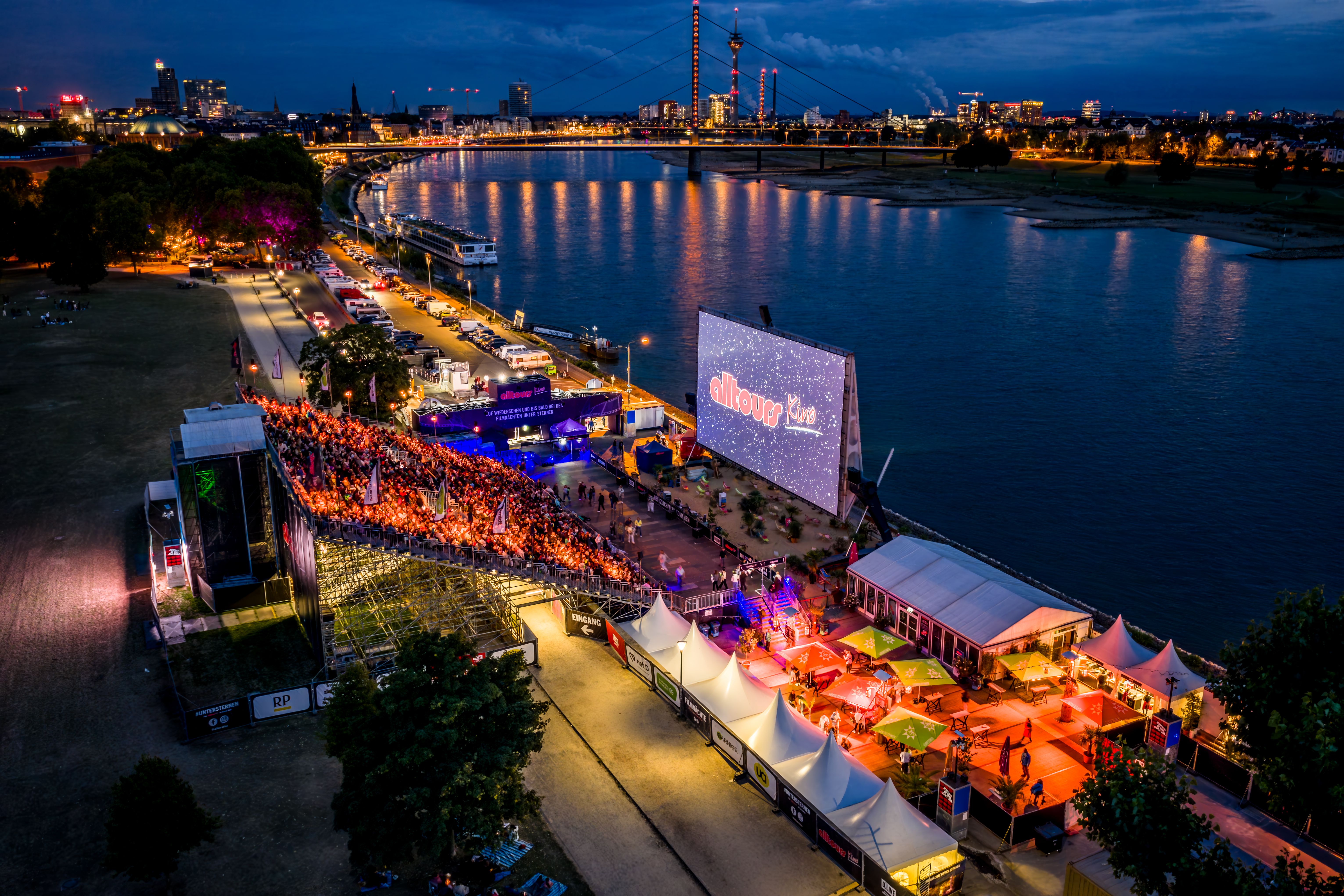 Luftaufnahme des alltours Kinos im Düsseldorfer Rheinpark bei Nacht mit großer Leinwand, gefüllter Zuschauertribüne, Rheinufer, Oberkasseler Brücke und beleuchteter Stadtsilhouette.