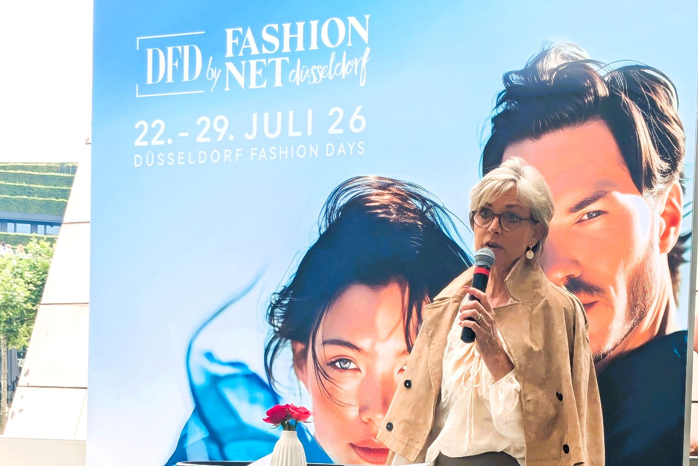 Ulrike Kähler spricht in ein Mikrofon an einem schwarzen Stehpult mit Notizen und einer Rose, hinter ihr das blaue Kampagnenmotiv der DFD by Fashion Net mit den Daten 22. bis 29. Juli 26