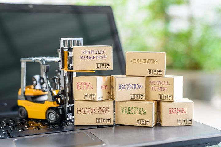 Miniatur-Gabelstapler vor einem Laptop stapelt Kartons mit den Aufschriften Aktien, Anleihen, ETFs, Rohstoffe, Fonds, Immobilienanlagen und Risiko.