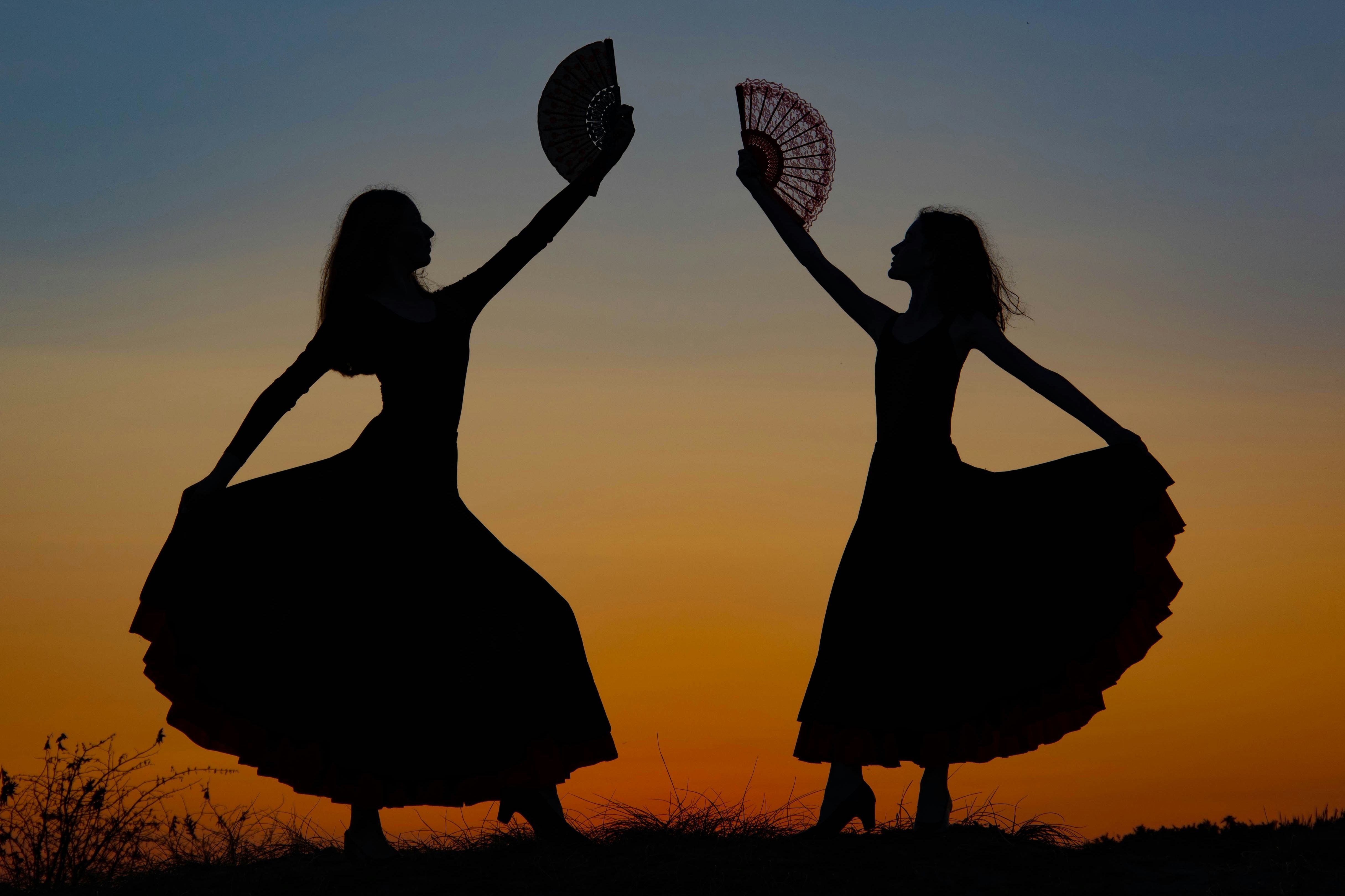 Silhouetten zweier Flamencotänzerinnen mit Fächern vor einem orangefarbenen Abendhimmel.
