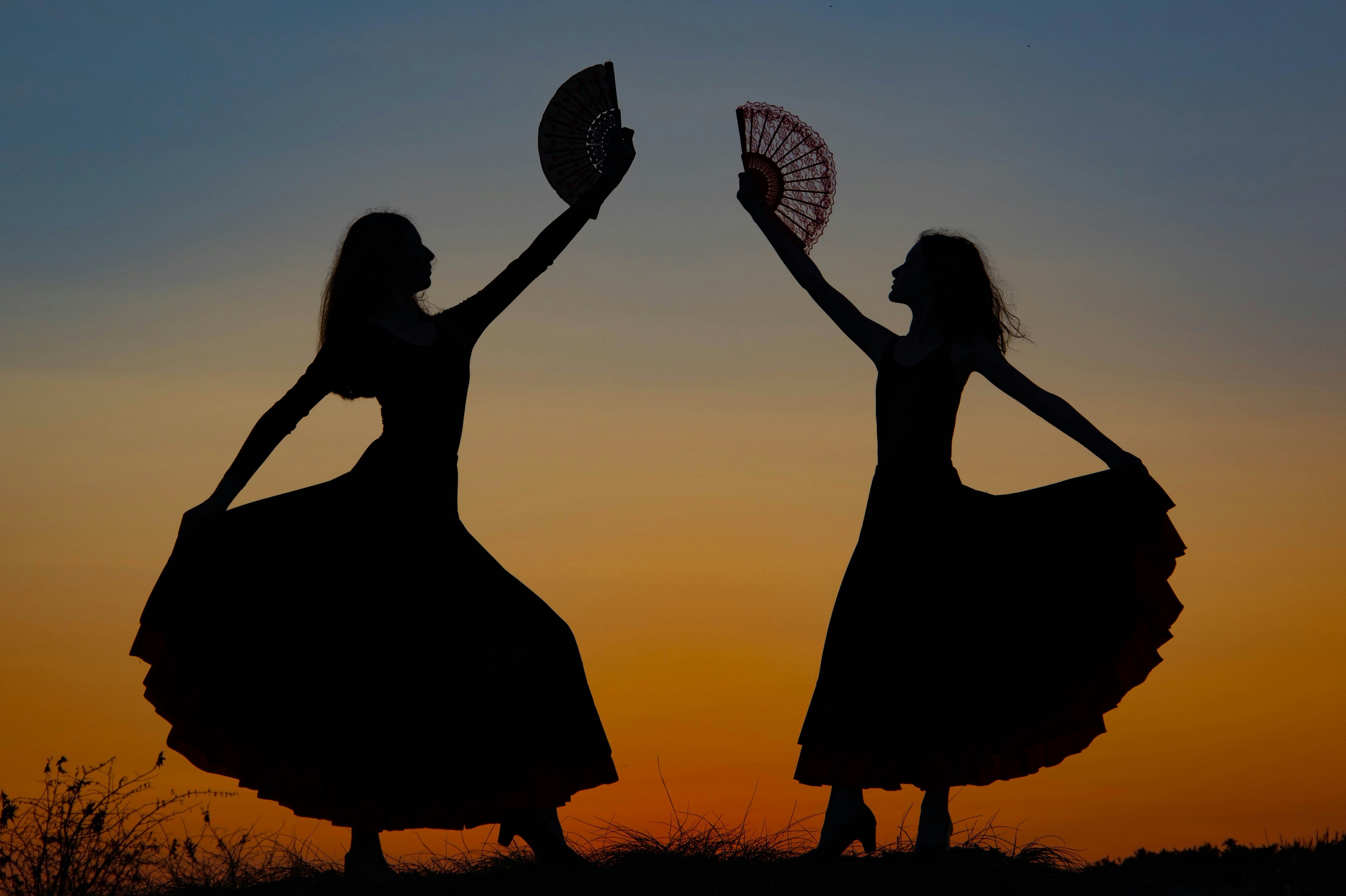 Silhouetten von zwei Flamenco-Tänzerinnen mit Fächern vor einem orangefarbenen Abendhimmel.