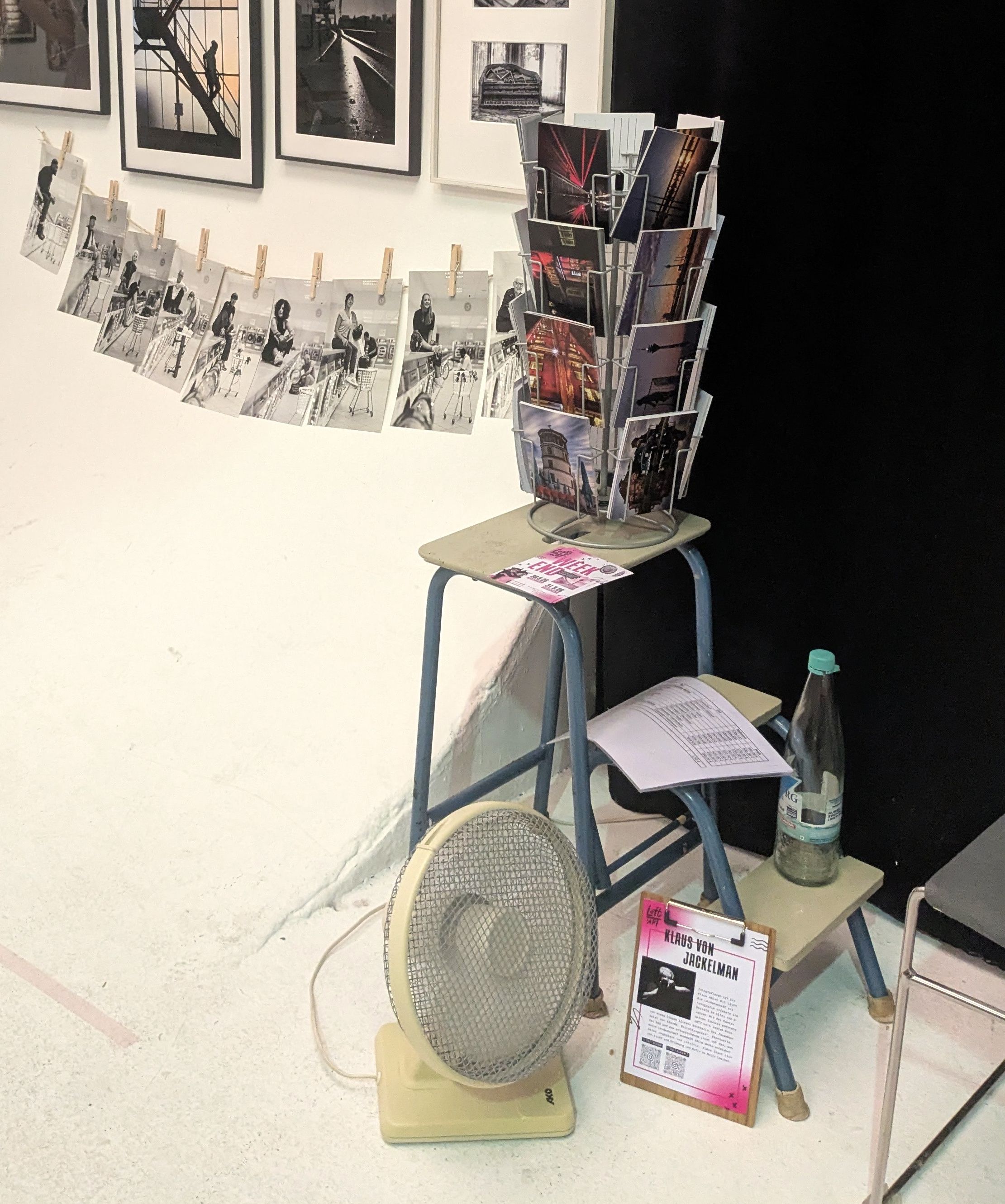 An einer weißen Wand hängen gerahmte Fotografien und an einer Schnur Schwarz-Weiß-Porträts der Serie Wash Salon, davor ein kleiner Ventilator, ein Postkartenständer und eine Karte mit dem Namen Klaus von Jackelmann.