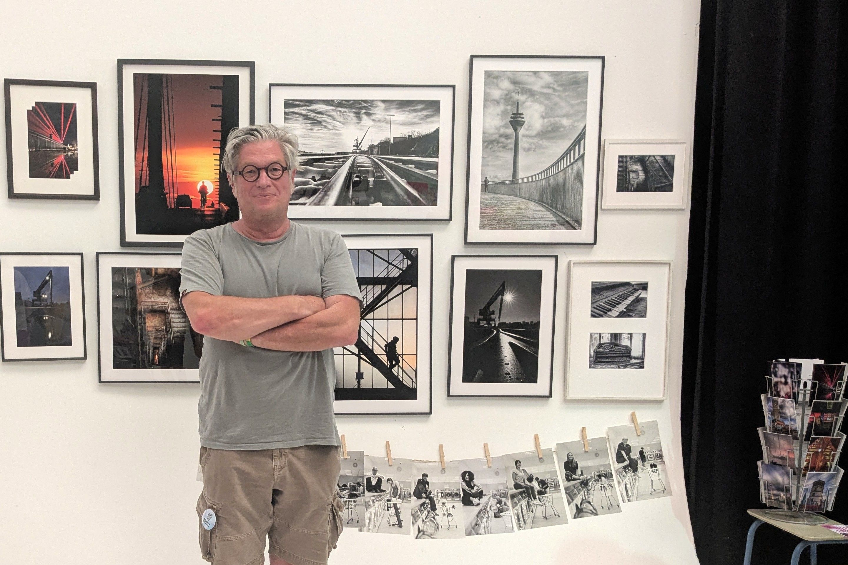 Ein Mann mit grauem T-Shirt und runder Brille steht mit verschränkten Armen vor einer Wand mit gerahmten Schwarz-Weiß- und Farbfotografien von Düsseldorf.