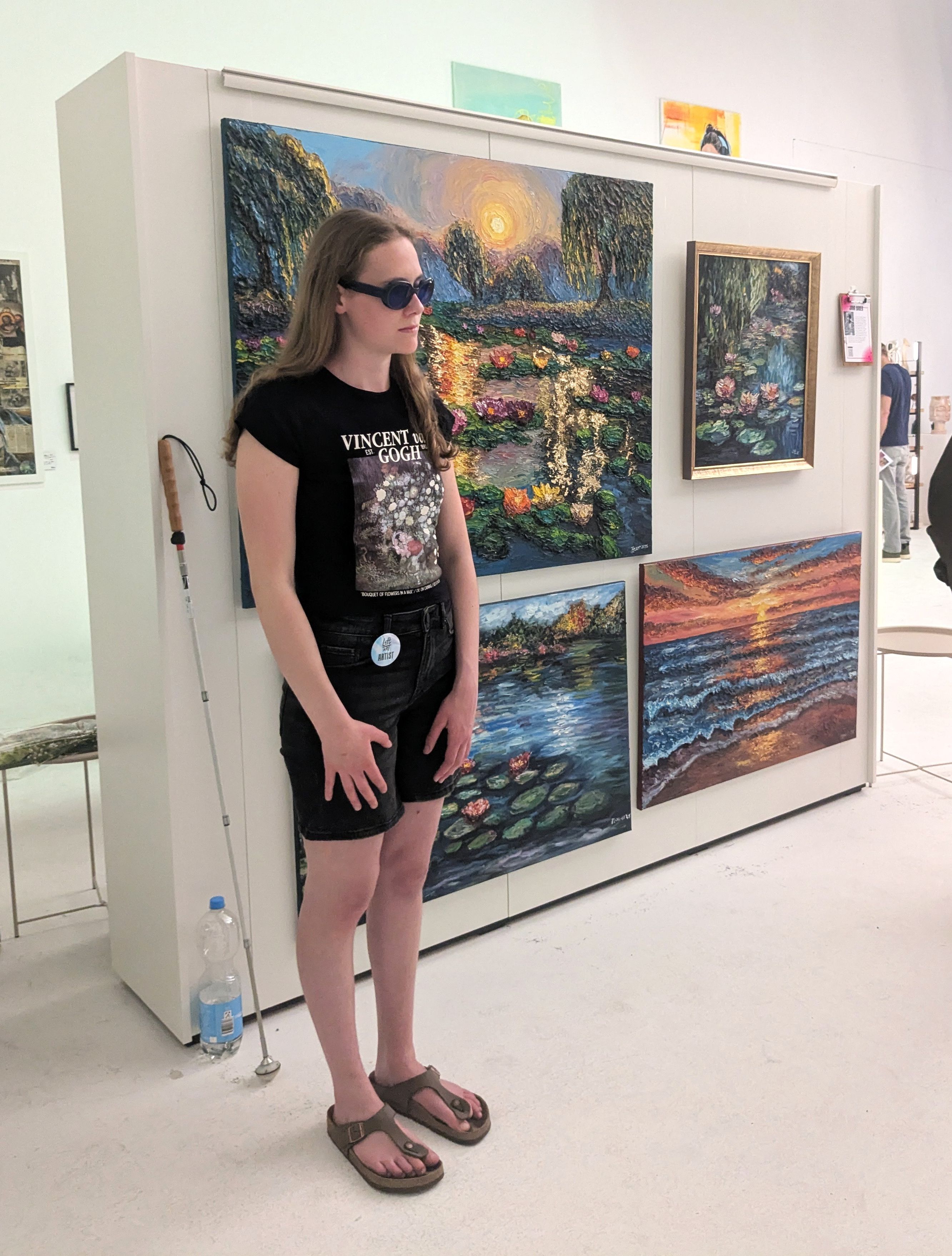 Eine junge Frau mit dunkler Sonnenbrille steht vor mehreren pastosen Landschaftsgemälden, links neben ihr lehnt ein weißer Blindenstock an der Wand.