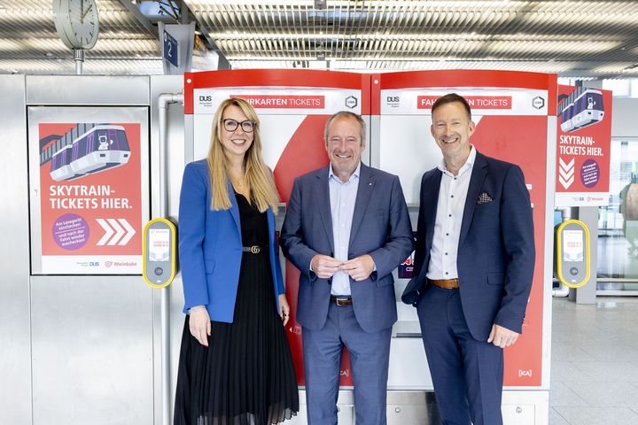Drei Vertreter von Rheinbahn, VRR und Flughafen Düsseldorf stehen am Flughafen vor den SkyTrain-Ticketgeräten und präsentieren das neue CALO-SkyTrain-Ticket.
