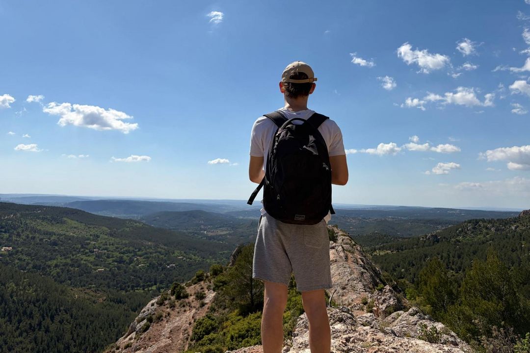 Victor steht auf einem Berg mit Rucksack steht mit dem Rücken zur Kamera vor einer weiten Aussicht