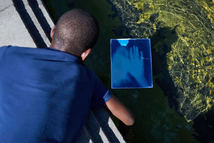 Dialog und Fotografie im KIT Düsseldorf Ein junger Teilnehmer hält während eines Fotografie-Workshops eine Cyanotypie über eine Wasserfläche.