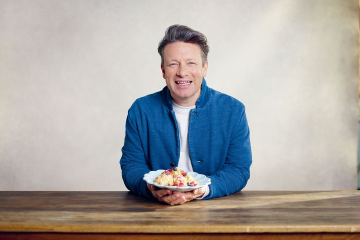 Jamie Oliver sitzt in blauer Jacke an einem Holztisch und hält einen Teller in den Händen.