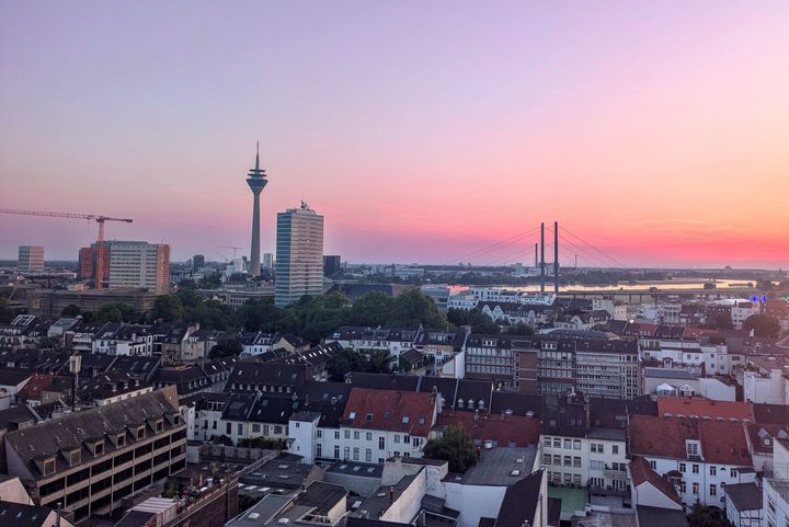 Blick über die Düsseldorfer Innenstadt bei Sonnenuntergang: dichte Wohnbebauung im Vordergrund, Rheinturm und Bürogebäude in der Mitte, Rheinkniebrücke und Rhein im Hintergrund, Baukran links im Bild.
