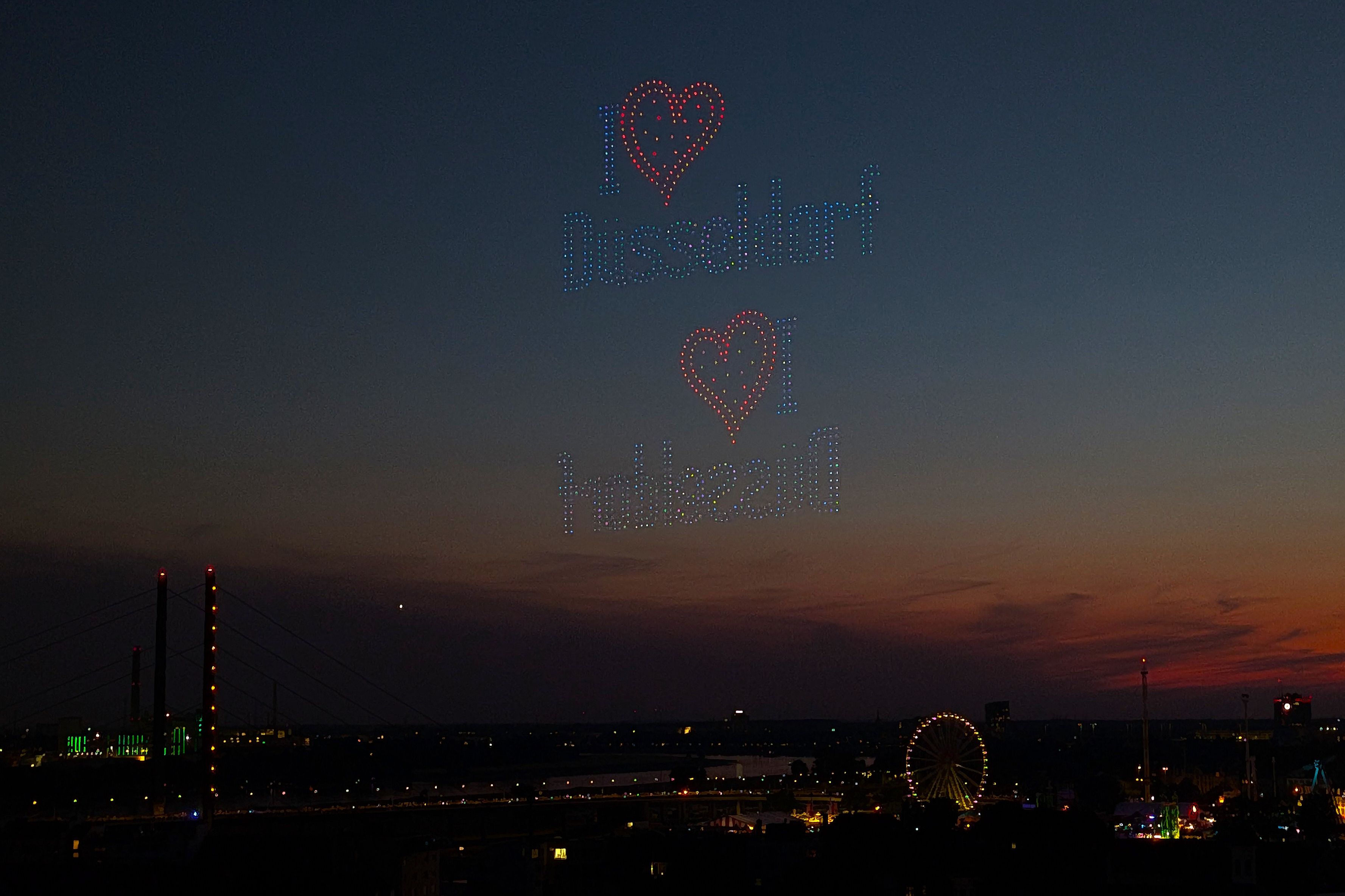 Drohnenshow über Düsseldorf mit zwei Herzen und dem Schriftzug „I love Düsseldorf“. Darunter sind die beleuchtete Rheinkirmes und das Riesenrad auf den Rheinwiesen zu sehen.