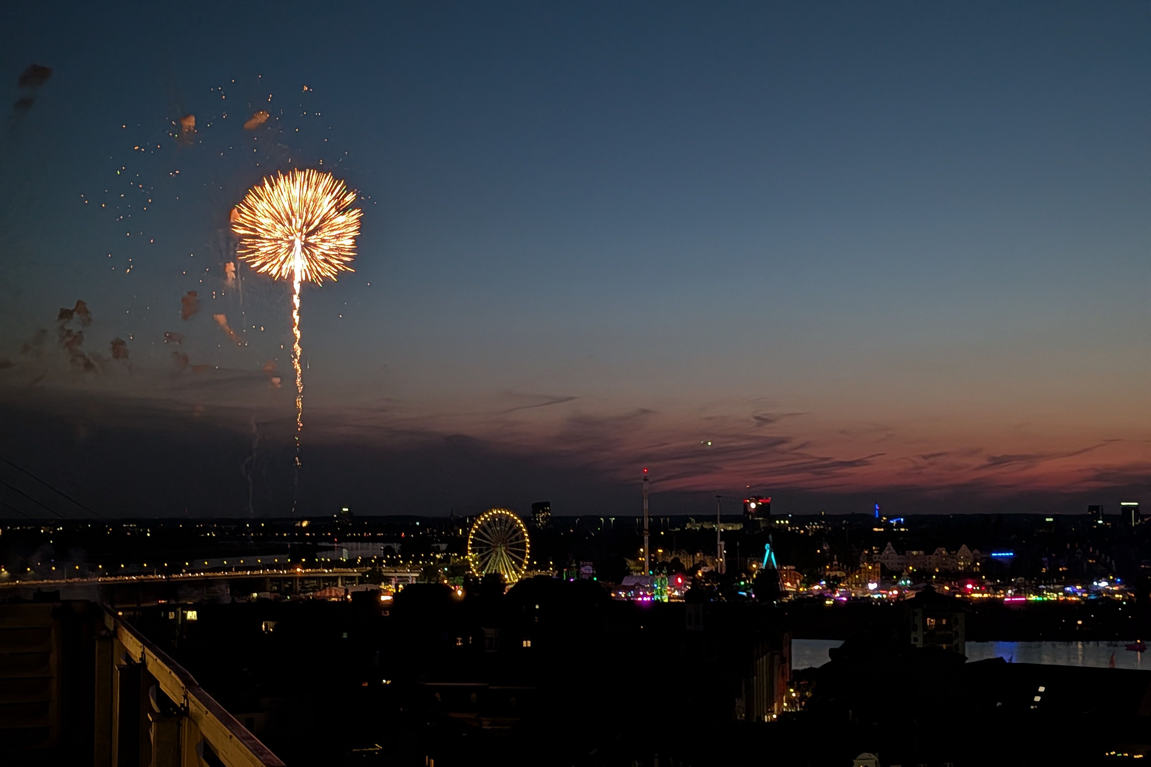 Feuerwerk über der Düsseldorfer Rheinkirmes bei Abenddämmerung. Im Hintergrund sind das Riesenrad, die Rheinwiesen und die Lichter der Kirmes zu sehen.