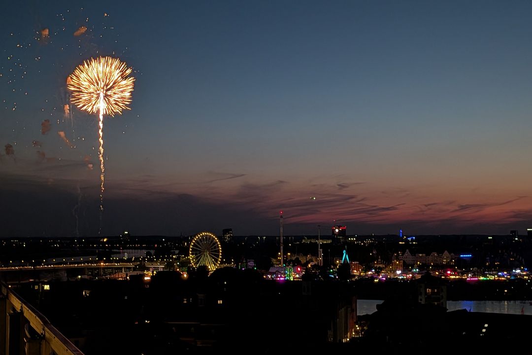 Blick über Düsseldorf auf die beleuchtete Rheinkirmes auf den Oberkasseler Rheinwiesen. Links steigt ein Feuerwerk in den Abendhimmel, darunter sind das Riesenrad und weitere Fahrgeschäfte zu sehen.