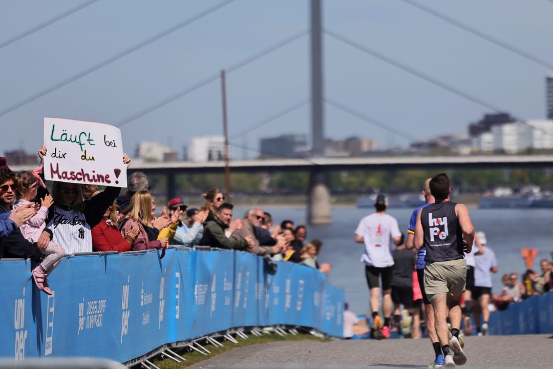 Zuschauerin hält Schild „Läuft bei dir du Maschine!" beim Uniper Düsseldorf Marathon 2026 am Rheinufer mit Oberkasseler Brücke im Hintergrund