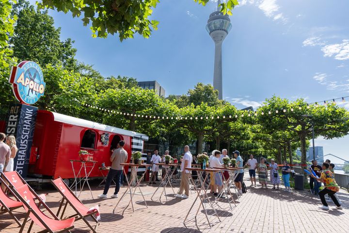 Sonnenterrasse des Apollo Rhein Rondells in Düsseldorf mit rotem Ausschankwagen, Gästen und Blick auf den Rheinturm