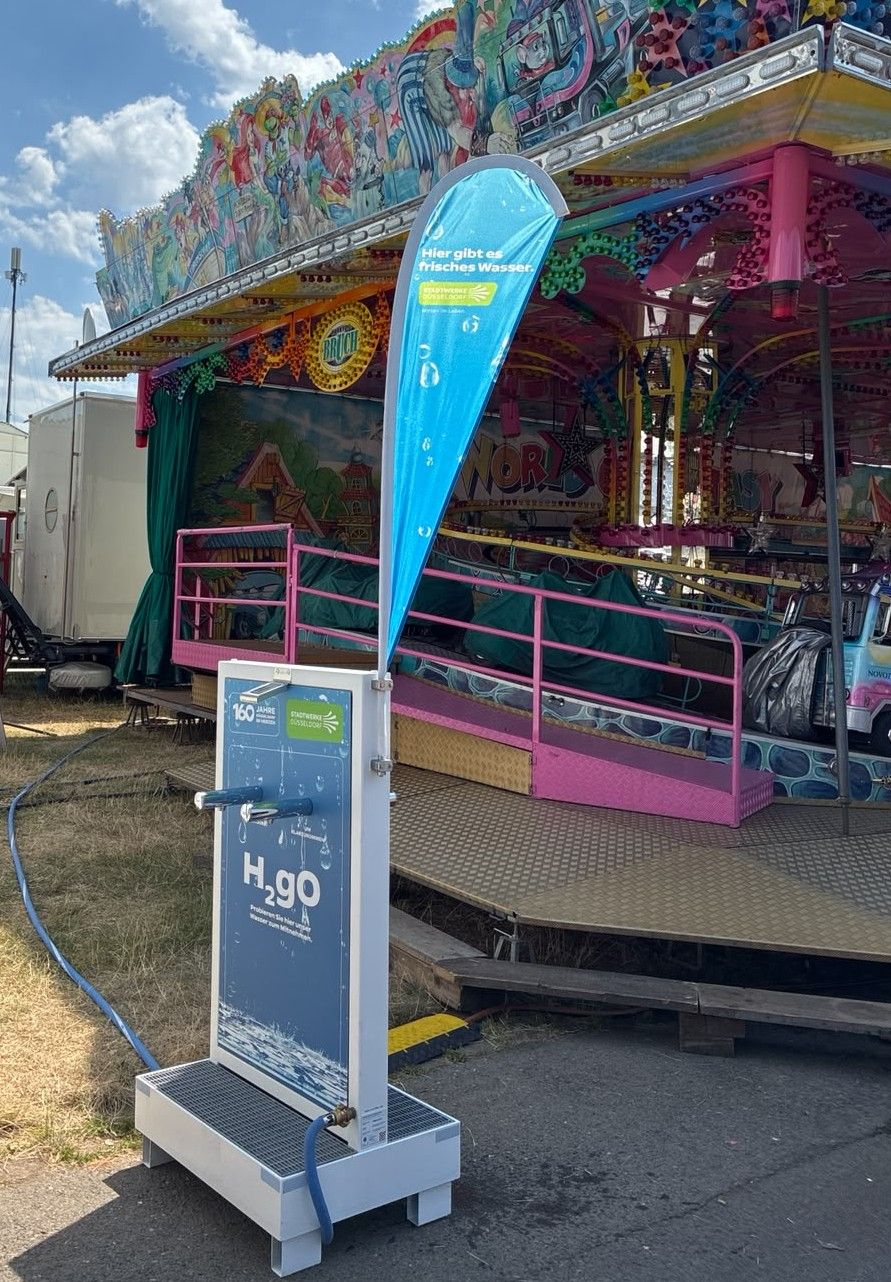 Eine kostenlose Trinkwasserstele der Stadtwerke Düsseldorf steht auf dem Gelände der Rheinkirmes vor einem bunt beleuchteten Fahrgeschäft.