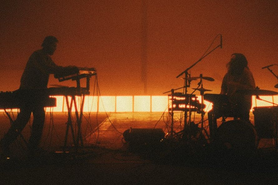 Zwei Musiker von Polynation als Silhouetten auf einer Bühne, einer am Synthesizer, einer am Schlagzeug, warmes orangefarbenes Gegenlicht
