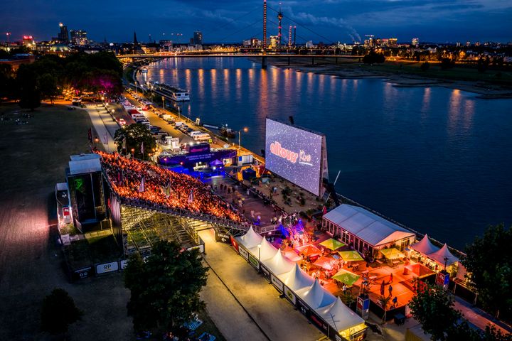 Blick von oben auf das alltours Kino im Rheinpark Düsseldorf bei Nacht mit großer Leinwand, Publikum, Rhein, Oberkasseler Brücke und beleuchteter Skyline.