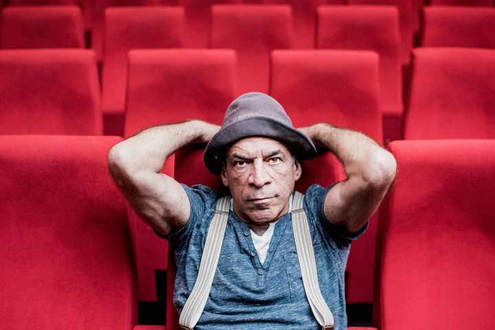 Peter Shub sitzt in einem roten Theatersaal, trägt Hut und Hosenträger und blickt frontal in die Kamera.