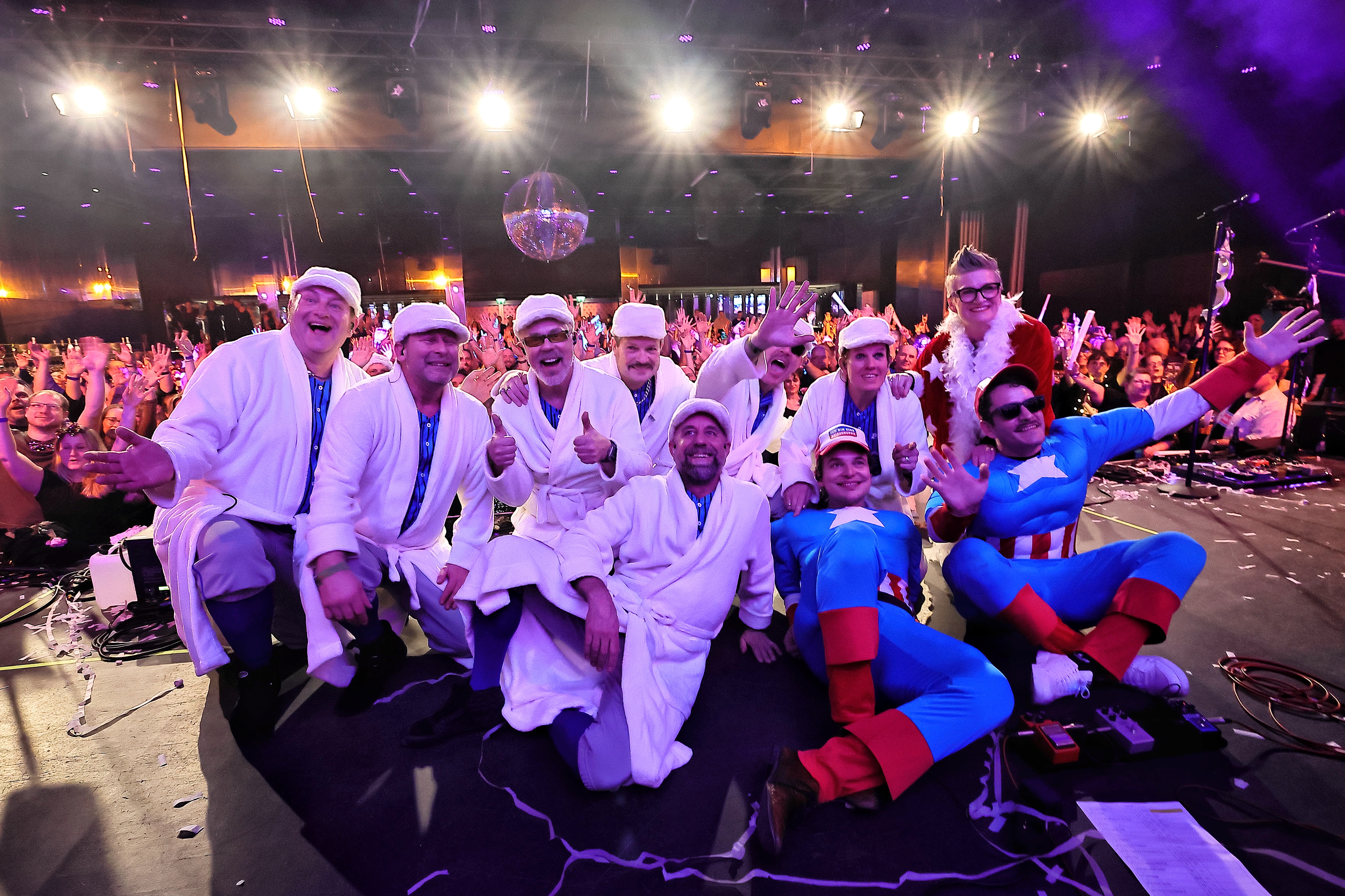 Kostümierte Musikergruppe posiert auf der Bühne, Publikum im Hintergrund.