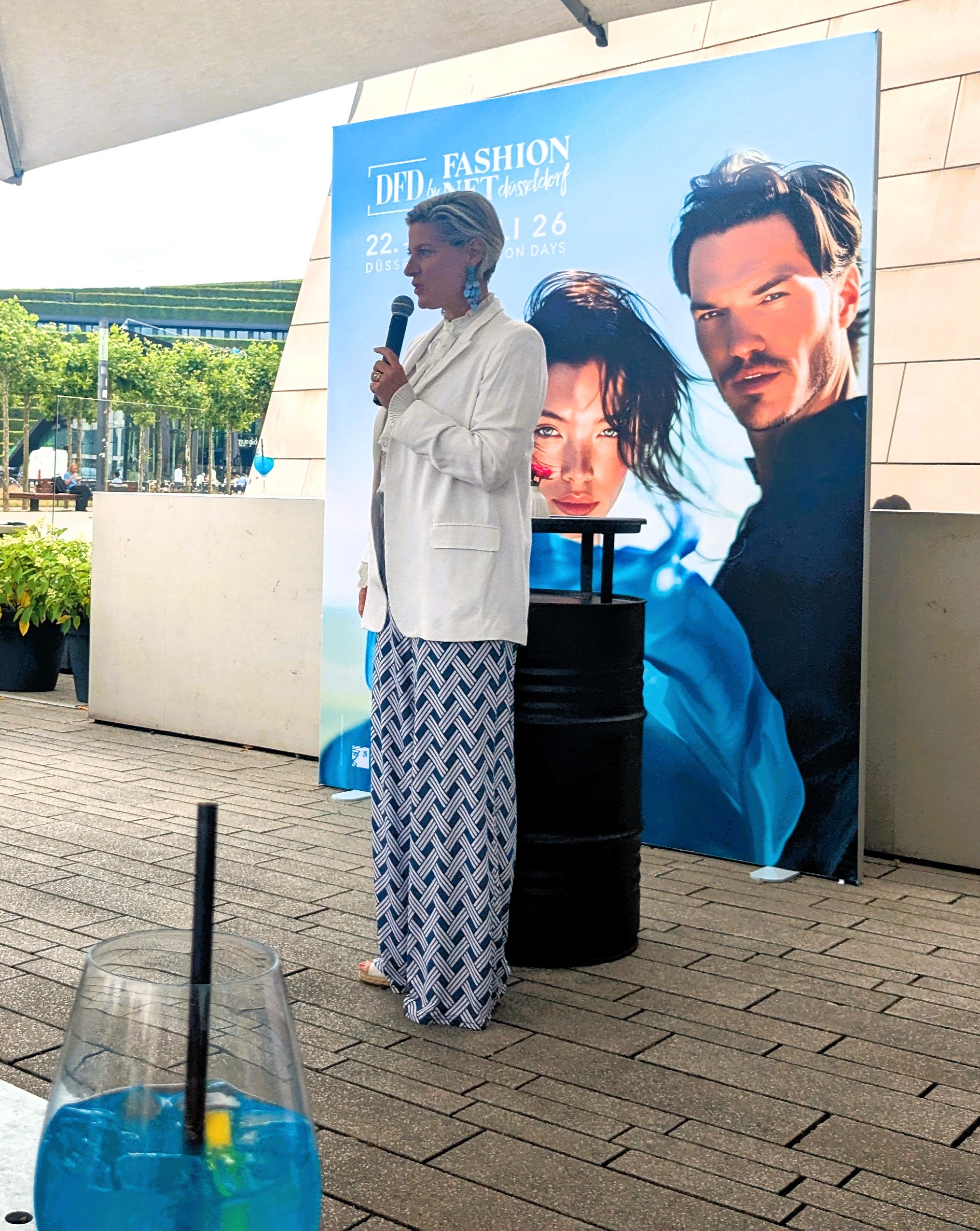 Ein blauer Drink mit Strohhalm im Vordergrund, dahinter spricht Andrea Greuner in weißem Blazer in ein Mikrofon, vor dem blauen Kampagnenmotiv der DFD by Fashion Net