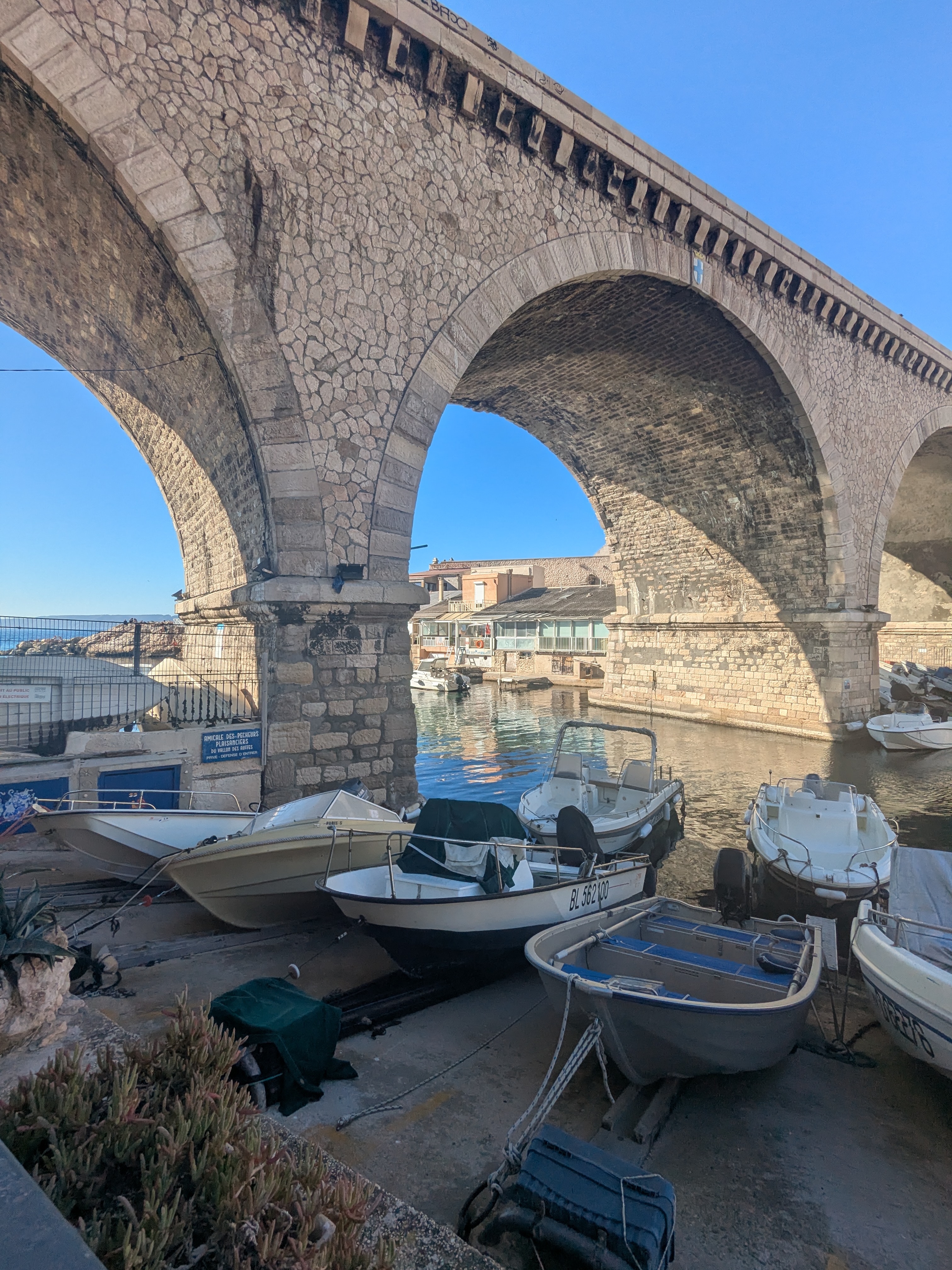 Blick aus dem Hafen des Vallon des Auffes durch die Steinbögen des Viadukts der Corniche Kennedy, mit festgemachten Fischerbooten im Vordergrund und blauem Himmel dahinter