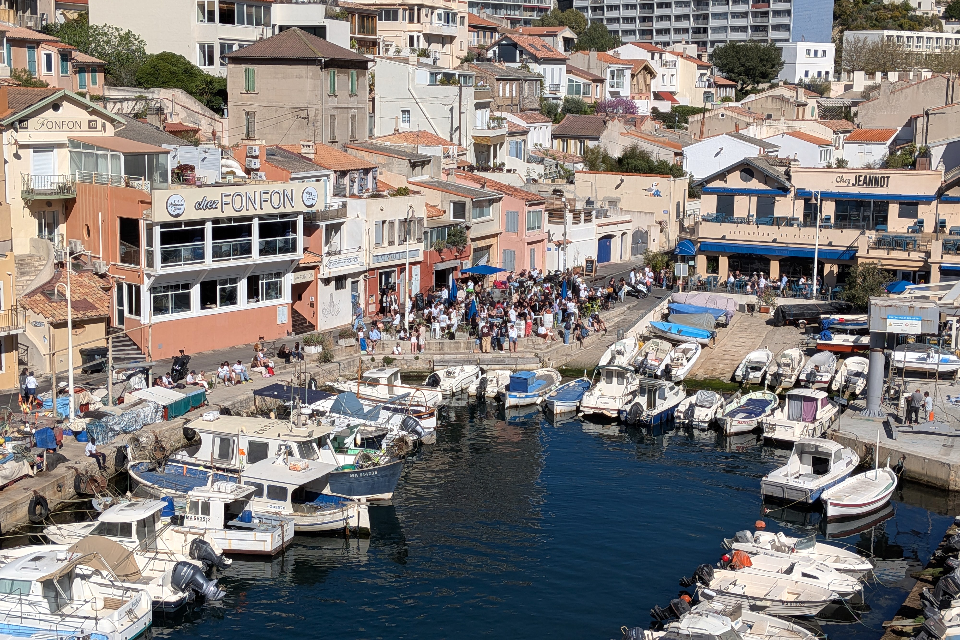 ogelperspektive auf den Vallon des Auffes in Marseille: Chez Fonfon links, Chez Jeannot rechts, Fischerboote im Hafen, Menschenmenge auf dem Kai an Ostern